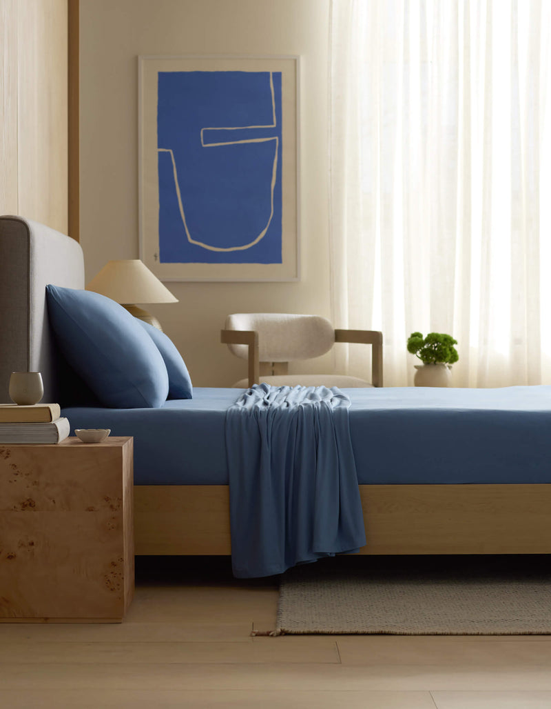 A modern bedroom features a bed with Cozy Earth’s Bamboo Jersey Sheet Set in blue, a light wood nightstand with a cup, books, and lamp, a chair, a plant, sheer curtains, abstract blue wall art, and soft natural light.