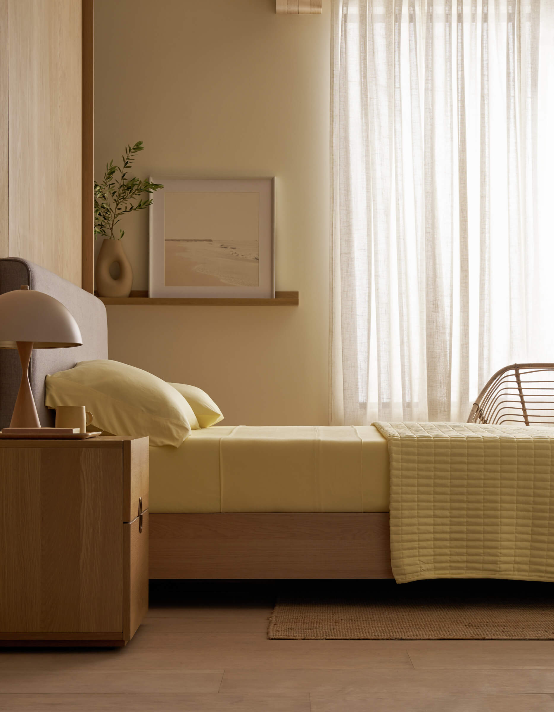 A minimal bedroom features a single bed with Cozy Earth’s Bamboo Jersey Flat Sheet in light yellow, a wooden nightstand holding a lamp and vase, a framed picture on a ledge, and sheer curtains letting in soft daylight.