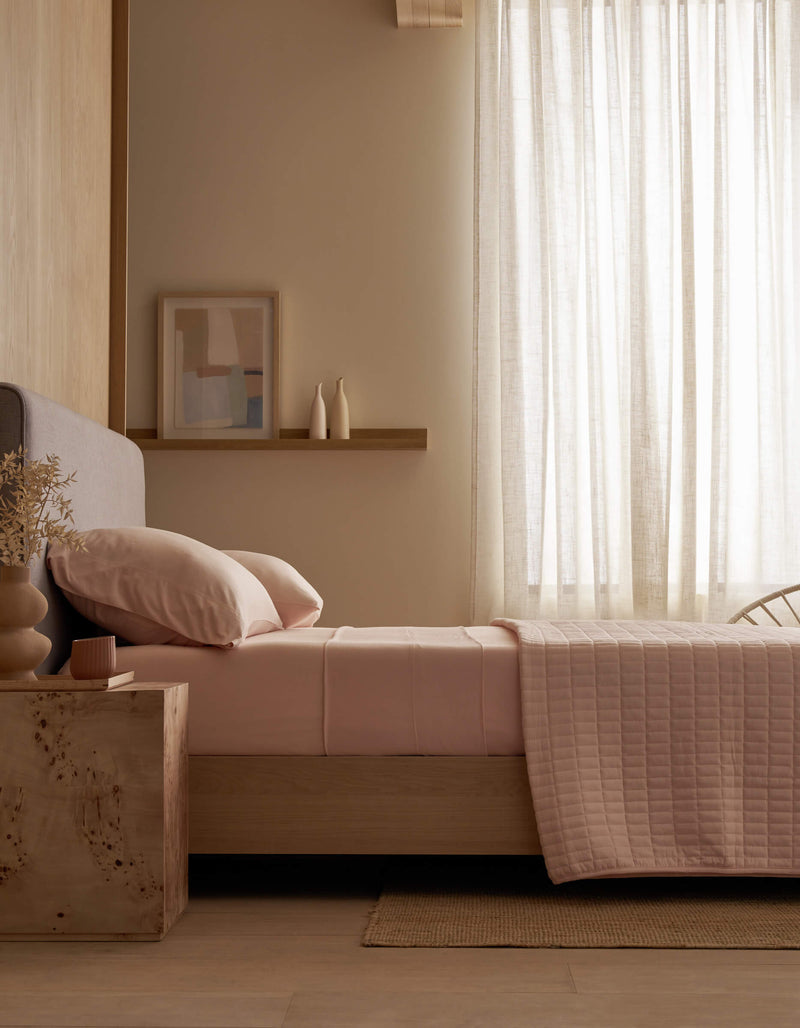 A minimalist bedroom features a neatly made bed with Cozy Earth Bamboo Jersey Flat Sheet in soft pink, a wooden nightstand holding a vase and candle, light wood walls, sheer curtains allowing natural light, and abstract art on a shelf.
