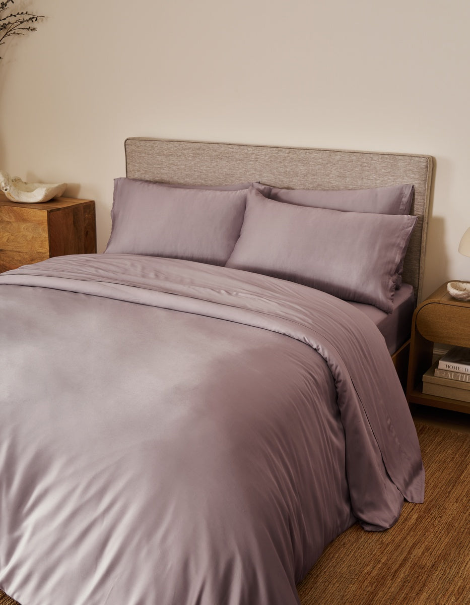 A neatly made bed features Cozy Earth’s Bamboo Bedding Ultimate Bundle in a light purple hue, complemented by two plush pillows and a cushioned headboard. Wooden nightstands with stylish decor and a small lamp are positioned on either side, enhancing the room’s warm, minimalist aesthetic.