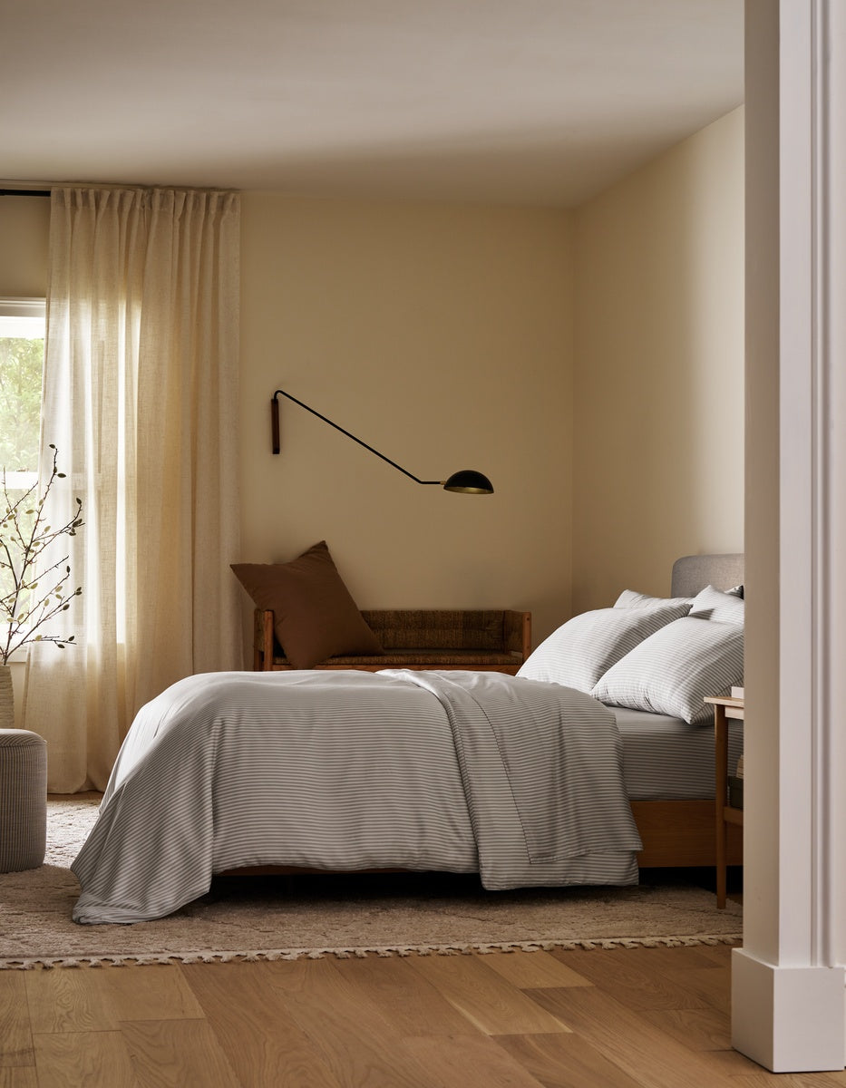 A cozy bedroom featuring a neatly made bed dressed in the Bamboo Bedding Ultimate Bundle by Cozy Earth, complemented by a brown pillow, a wall-mounted lamp, a wooden bench, beige curtains, and light wooden floors. Natural light filters through the window.