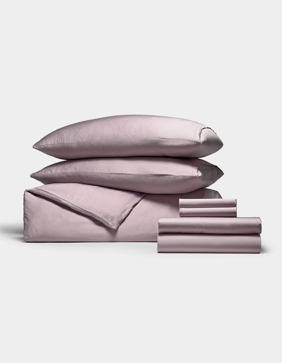 A neatly stacked set of Cozy Earth’s Bamboo Bedding Ultimate Bundle, featuring two pillows, a folded duvet, a flat sheet, and several smaller folded sheets in pale pink, all arranged against a plain white background.|Color:Lavender