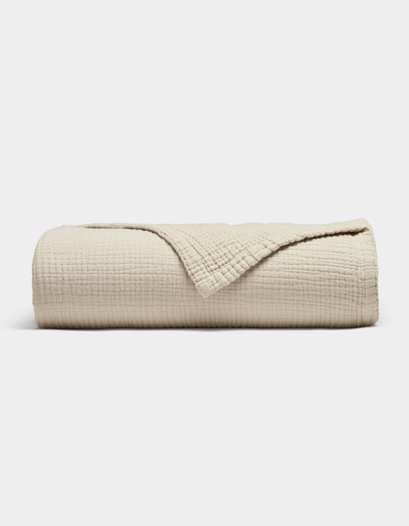 The Cozy Earth Cotton Gauze Blanket, in beige with a soft grid texture, is neatly rolled with a corner folded back to reveal its fabric, shown against a plain white background.