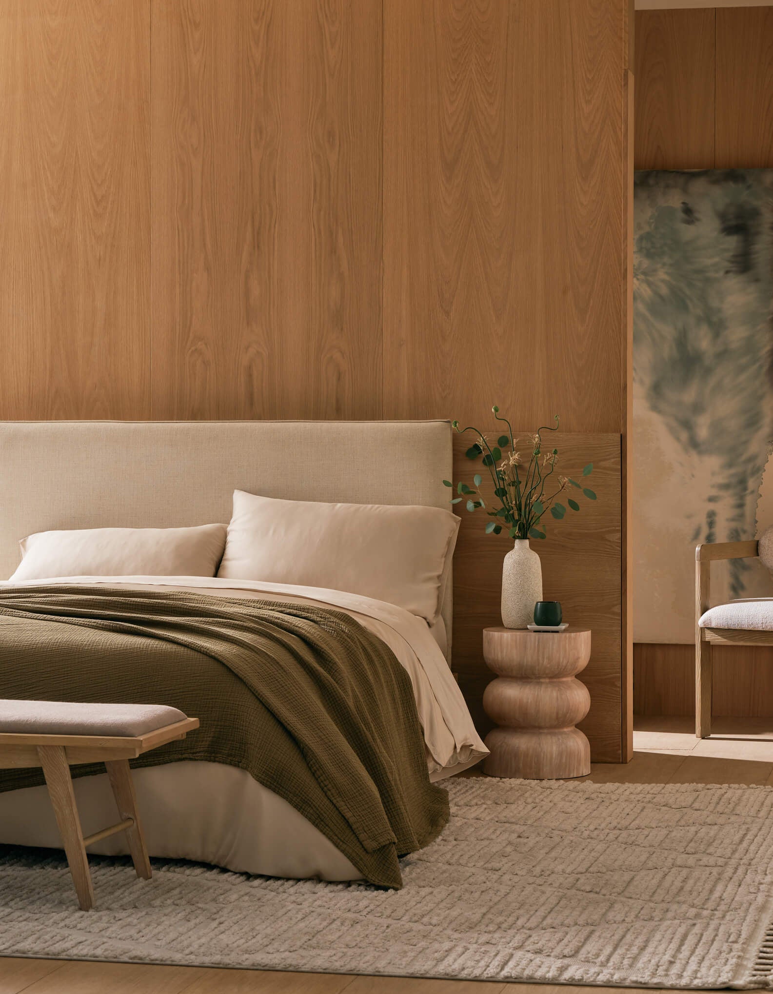 A modern, minimalist bedroom with a beige bed, Cozy Earth’s Cotton Gauze Blanket in olive green, light wooden walls, a textured rug, sculptural nightstand with a small vase of greenery, cushioned chair, and abstract wall art.