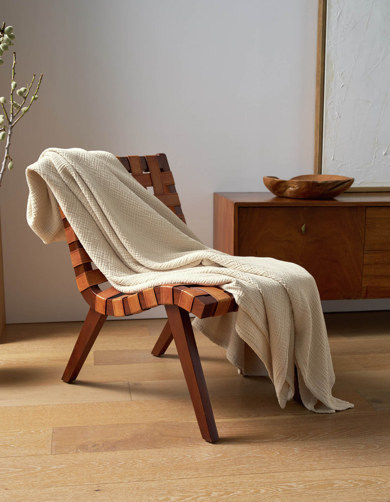 A Cozy Earth Cotton Gauze Blanket in beige is draped over a wooden woven chair beside a modern sideboard, accented by a decorative bowl and abstract wall art in a sunlit, minimalist room.