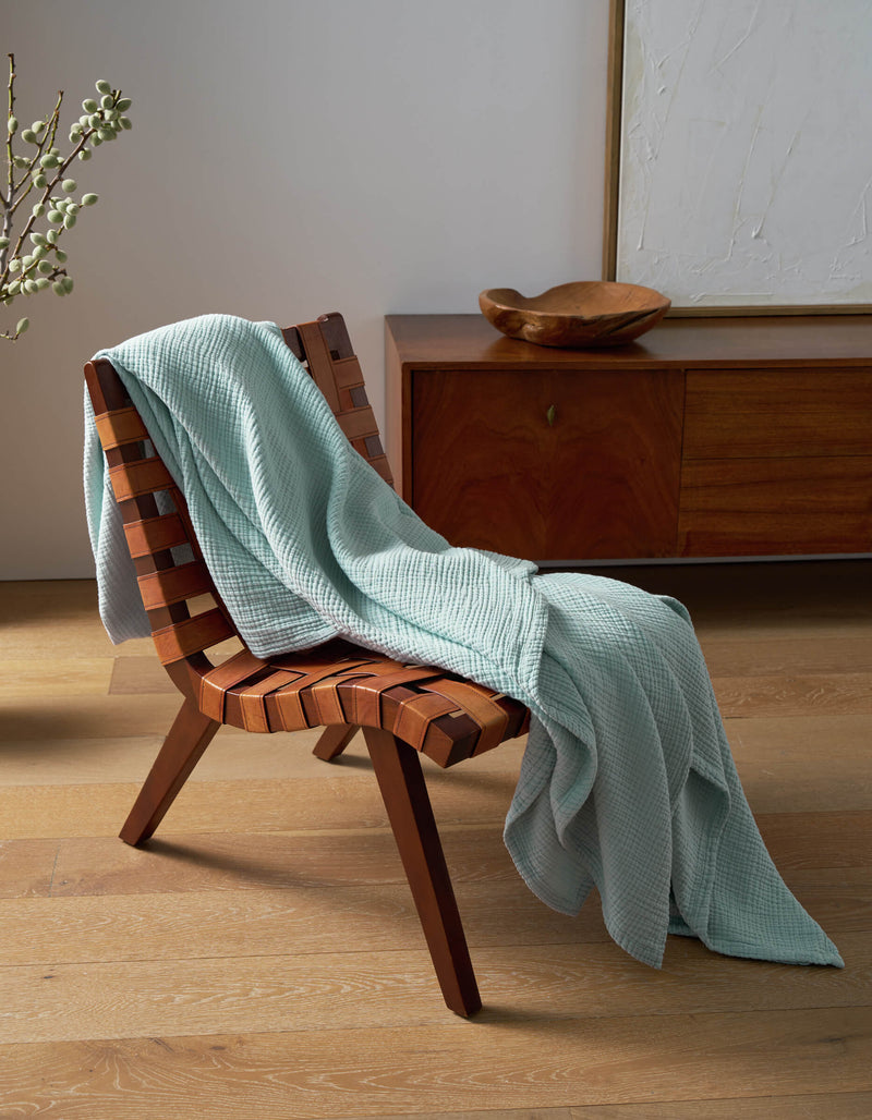 A wooden woven chair draped with the Cozy Earth Cotton Gauze Blanket in light blue sits on a wooden floor beside a sideboard topped with a decorative bowl and framed abstract art.