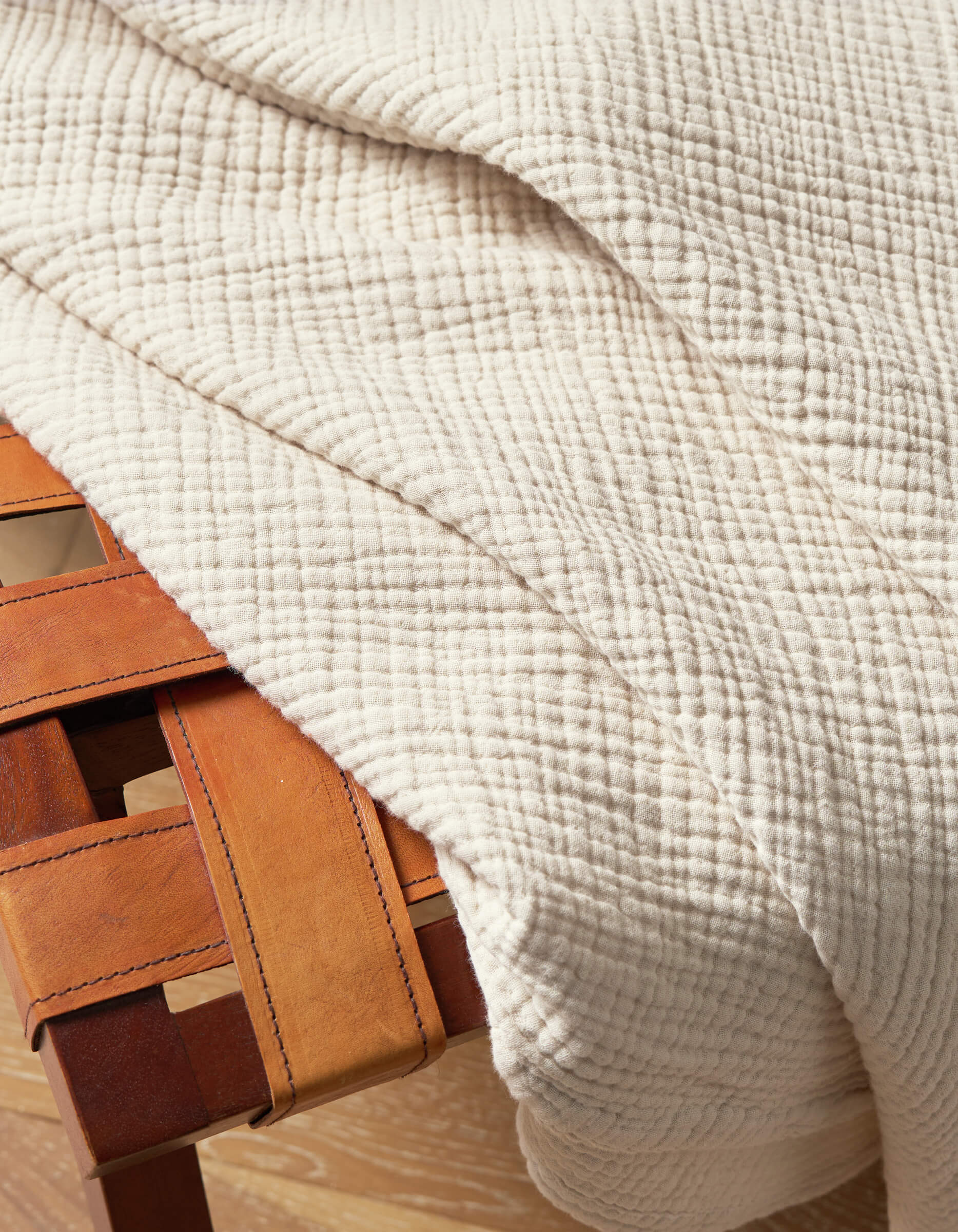 The Cozy Earth Cotton Gauze Blanket, in a cream color with a textured grid pattern, is draped over the corner of a woven leather bench featuring tan straps and a wooden frame.