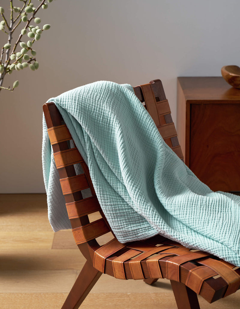 A Cozy Earth Cotton Gauze Blanket in light blue is draped over a woven wooden chair, next to a wooden side table with a vase of branches, all set on a light wood floor.