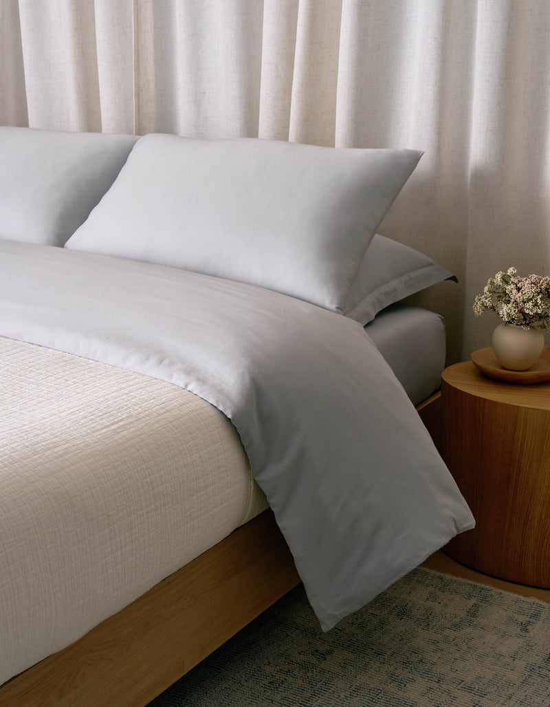A Waterfront Grid Matelasse Blanket by Cozy Earth in pale gray covers a neatly made bed with two pillows, beside a wooden nightstand holding a small vase of dried flowers, set against light-colored curtains.