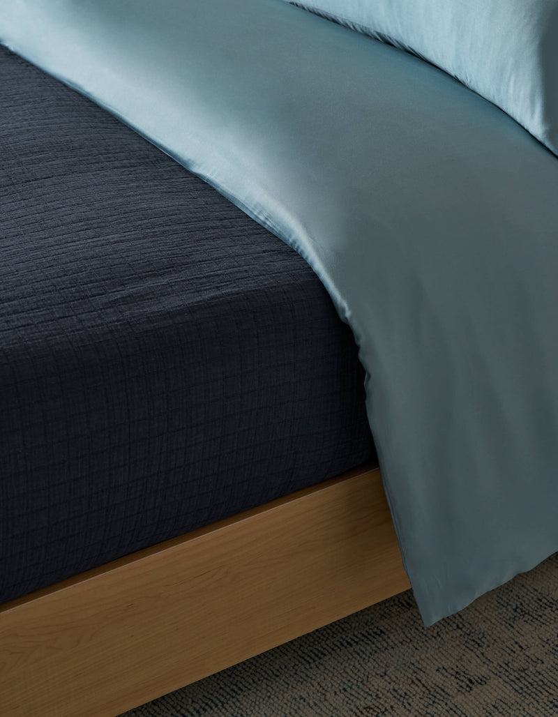 Close-up of a neatly made bed featuring the Cozy Earth Waterfront Grid Matelasse Blanket, a dark textured layer under a light blue satin comforter, on a wooden bed frame over a patterned carpet.