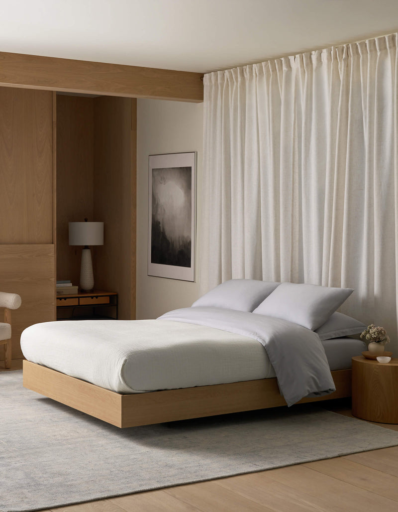 A modern, minimalist bedroom featuring a light wood platform bed, the Cozy Earth Waterfront Grid Matelasse Blanket in gray, a round wooden nightstand, lamp, abstract wall art, light curtains, and soft natural lighting.