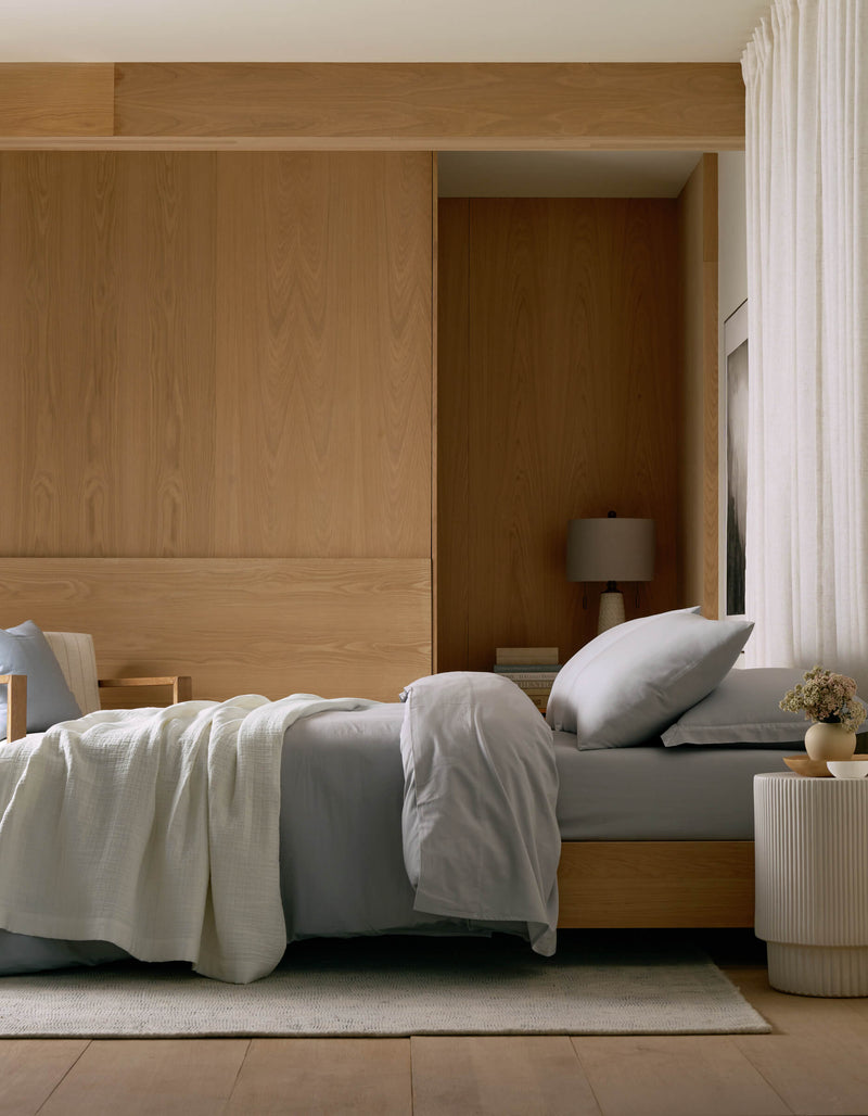 A modern, minimalist bedroom with a neatly made bed featuring Cozy Earth’s Waterfront Grid Matelasse Blanket in light gray, a wooden headboard, table lamp, round white side table with flowers, light wood paneling, and cream curtains.