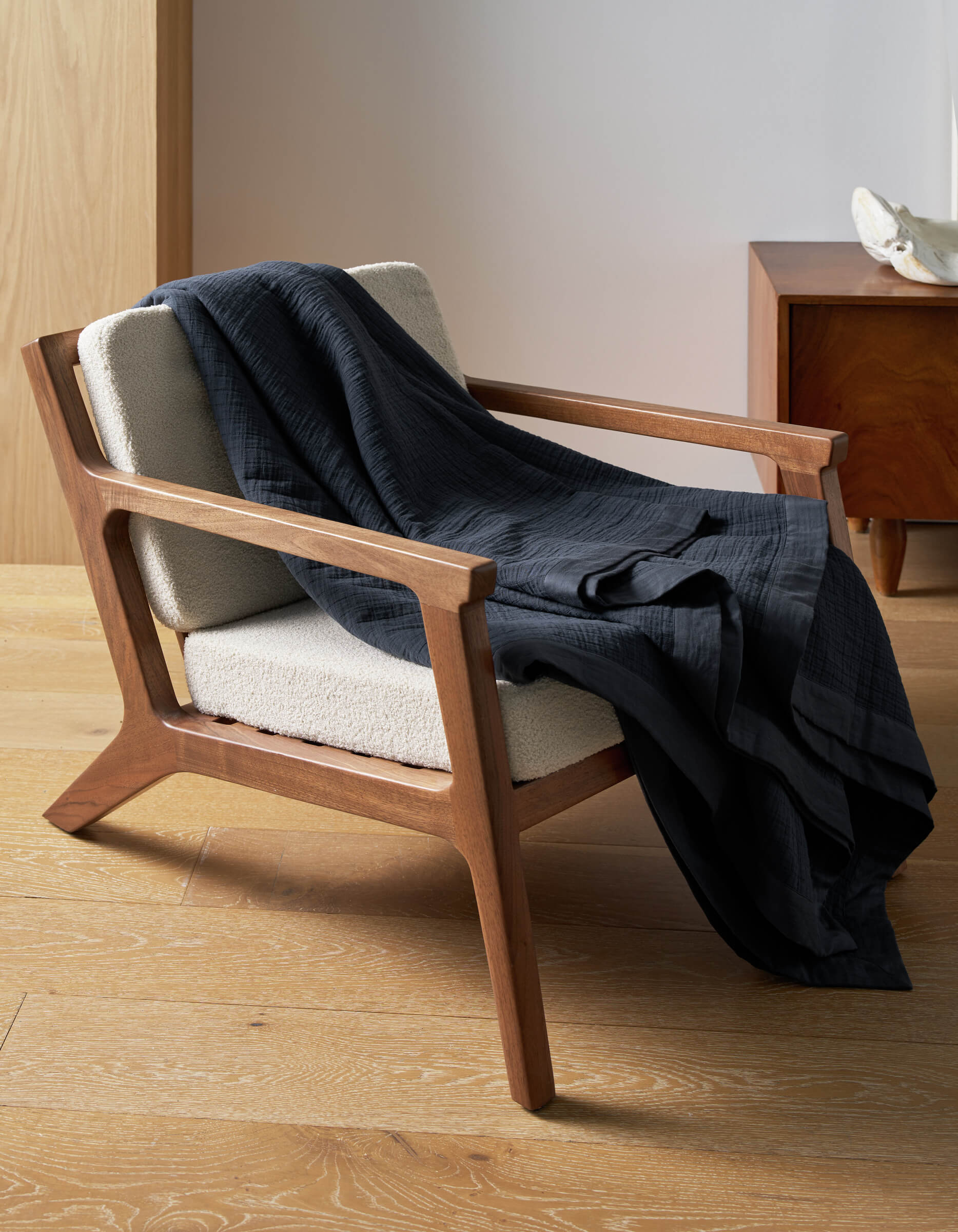 A wooden armchair with cream upholstery sits on a light wood floor, draped with a Cozy Earth Waterfront Grid Matelasse Blanket. A wooden side table and decorative object appear in the background.|Color:Eclipse