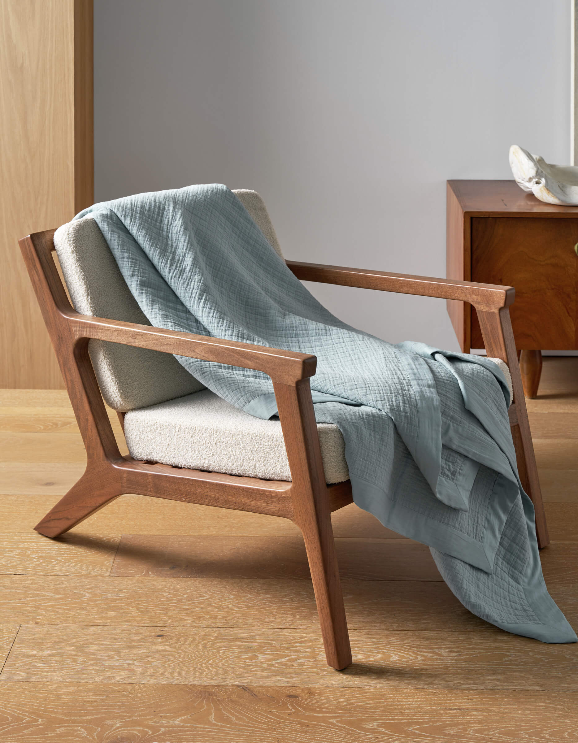 A mid-century modern wooden armchair with cream upholstery sits on a light wood floor, draped with the Cozy Earth Waterfront Grid Matelasse Blanket in light blue. In the background, a wooden nightstand and decorative object are visible.