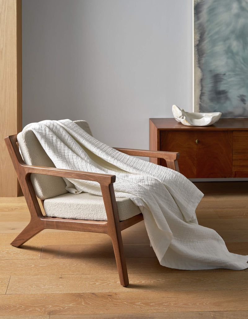 A wooden armchair with cream upholstery sits on a light wood floor, draped with the Cozy Earth Waterfront Grid Matelasse Blanket. In the background, there’s a wooden sideboard with a white bowl and large abstract artwork on the wall.