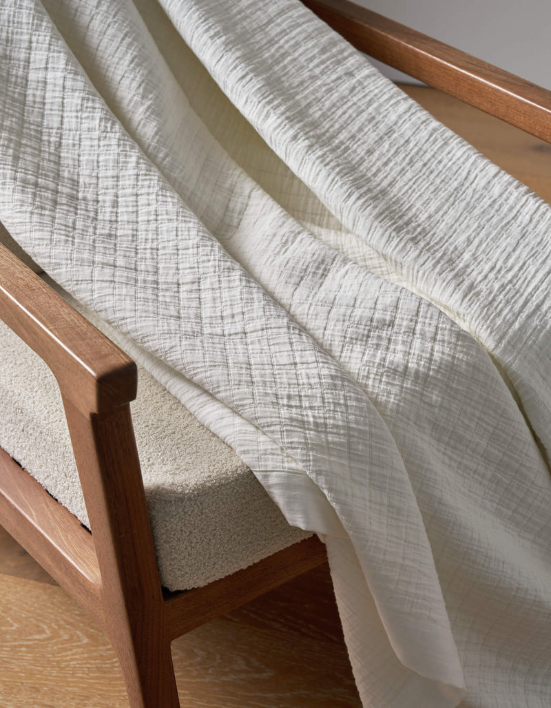 The Cozy Earth Waterfront Grid Matelasse Blanket, in a soft off-white with a subtle crinkled pattern, is draped over the arm of a wooden chair featuring a light cushioned seat on a wooden floor.