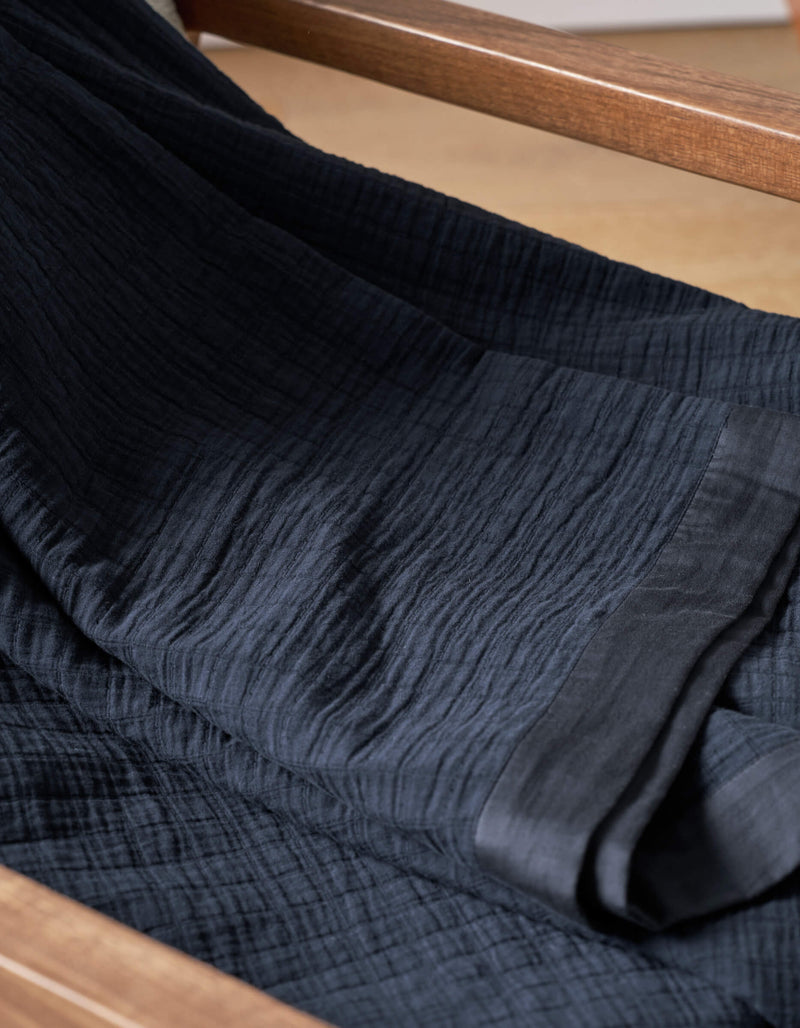 A close-up of Cozy Earth's Waterfront Grid Matelasse Blanket in dark navy, draped over wood, highlights its textured fabric with fine crinkles and a smooth edge.