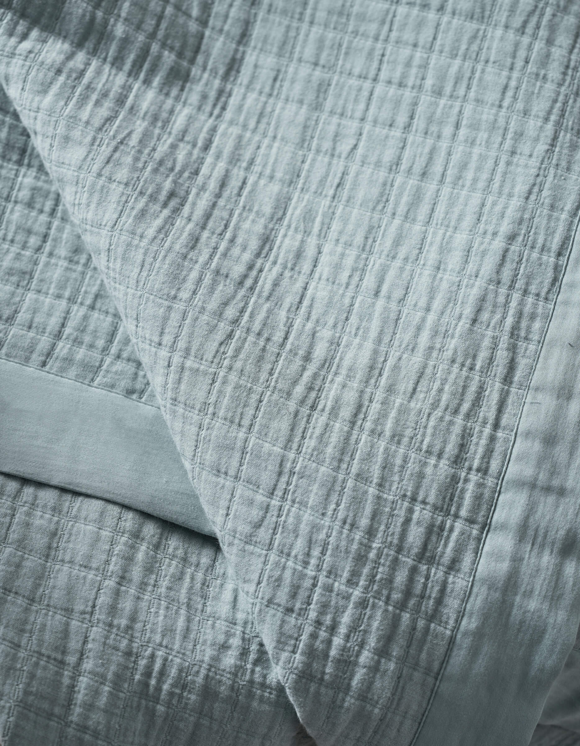 A close-up of the Cozy Earth Waterfront Grid Matelasse Blanket, light blue with a soft, textured grid pattern, neatly folded on a matching sheet. The fabric looks inviting and cozy.