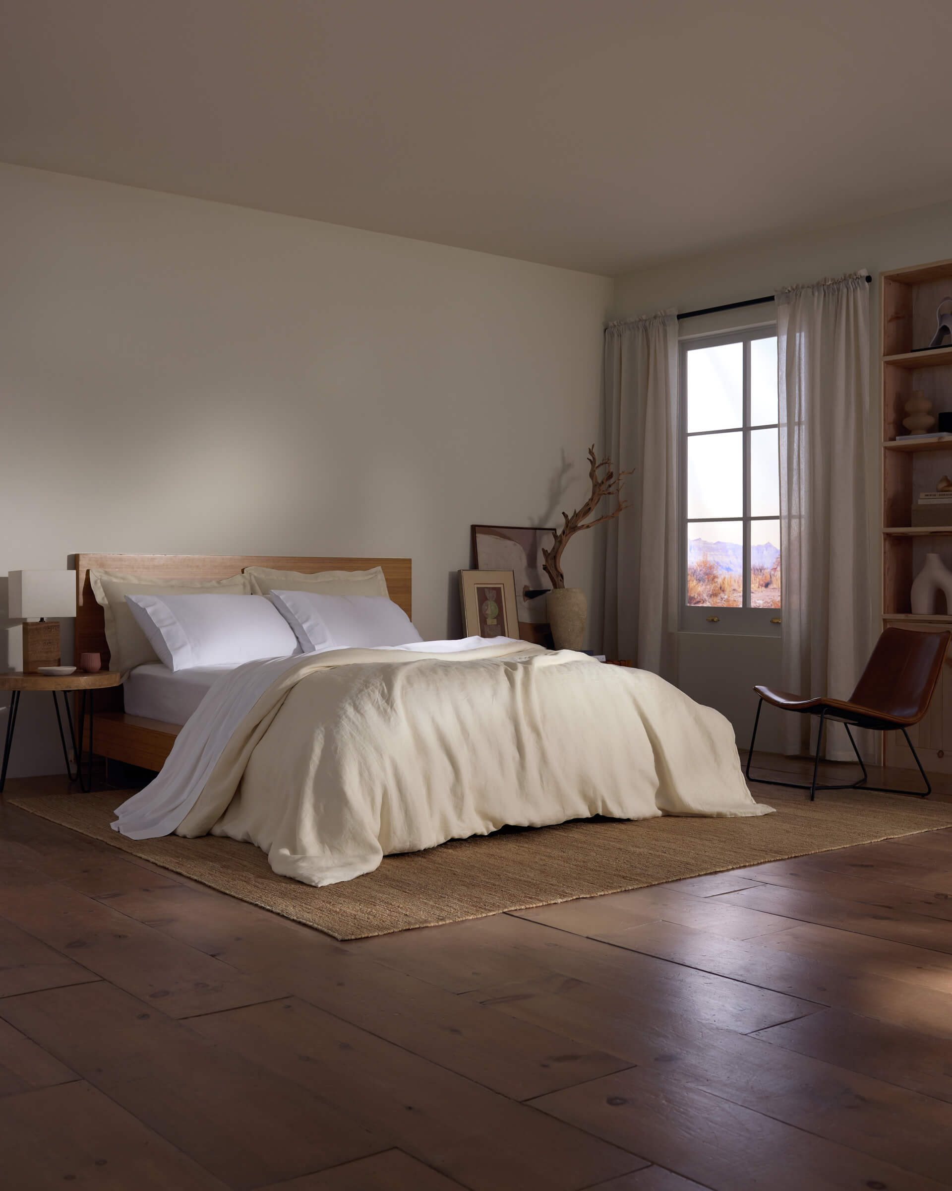 A minimal bedroom with wooden floors, a Cozy Earth Linen Duvet Cover on white bedding, a wooden bed frame, bedside table, window with sheer curtains, and a chair by shelves and decor on the right wall.|Color:Flax