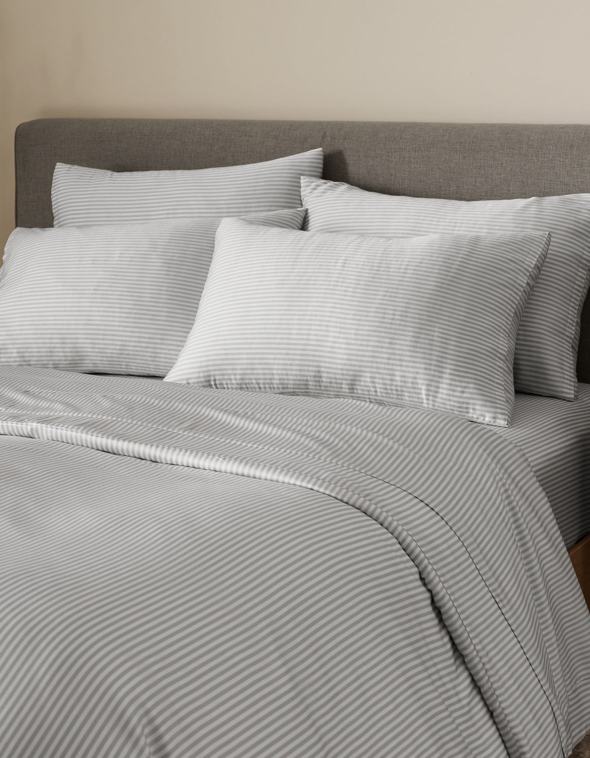 A neatly made bed with a gray upholstered headboard is dressed in light gray and white pinstriped bedding and comforter, with multiple pillows—including Bamboo Pillowcases by Cozy Earth—arranged against a beige wall.