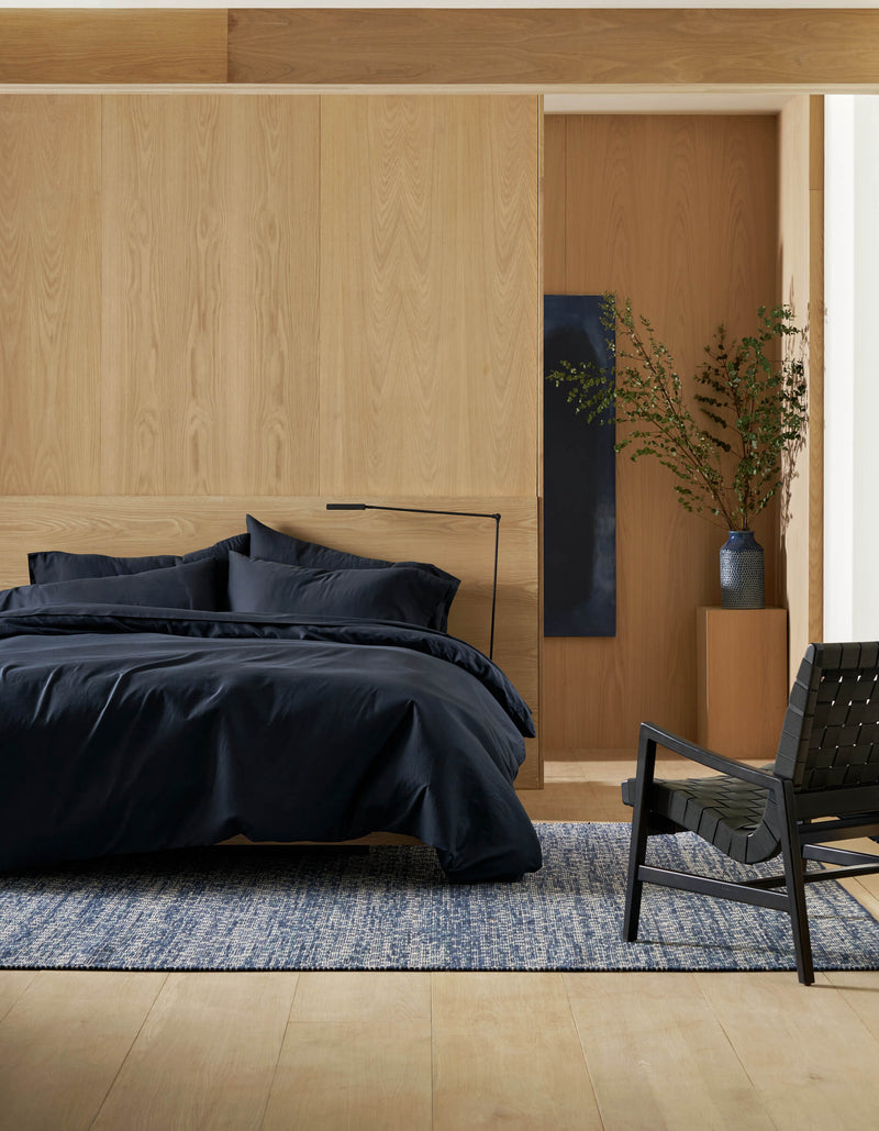 A modern bedroom with light wood walls, a bed featuring Cozy Earth's Soft-Wash Cotton Bedding Core Bundle, a blue and white rug, a black woven chair, and a tall vase with green branches on a wooden pedestal.