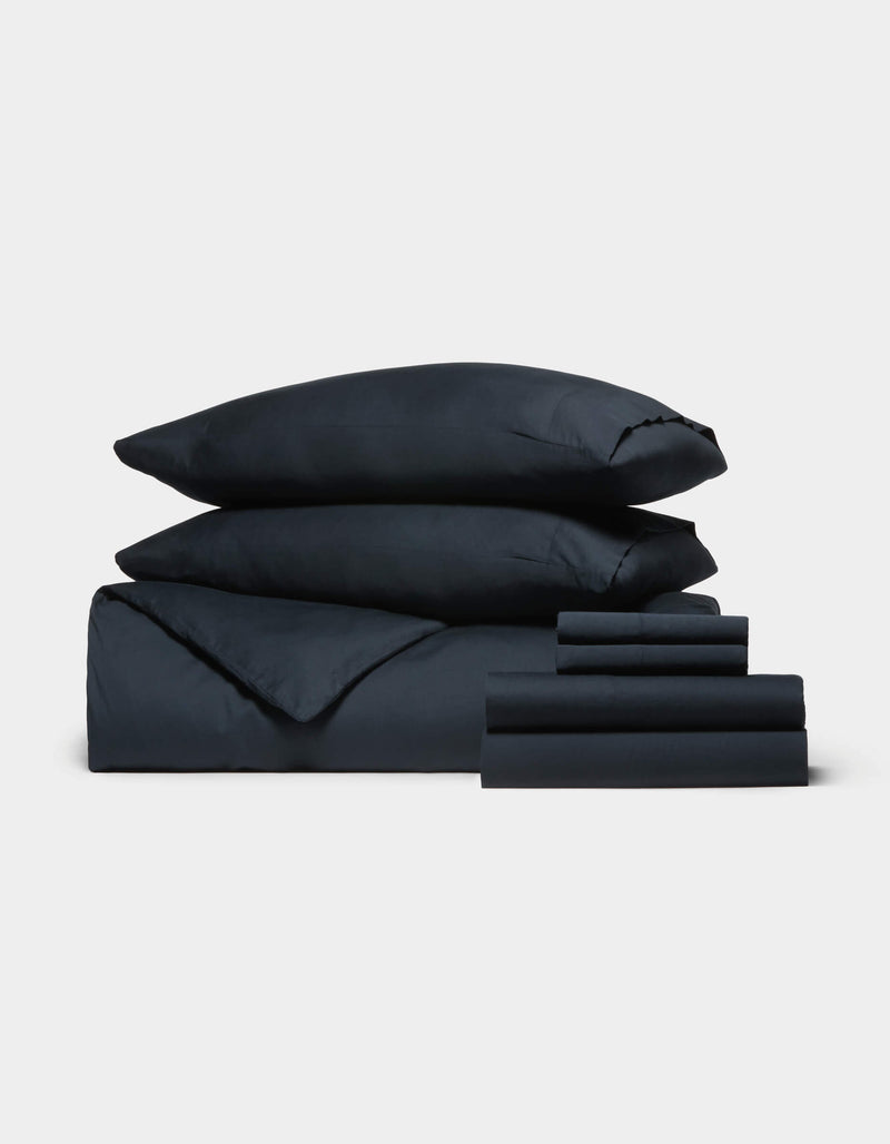 The Cozy Earth Soft-Wash Cotton Bedding Core Bundle, featuring two stacked pillows, a duvet, and several sheets in dark navy blue, is neatly arranged against a plain white background.