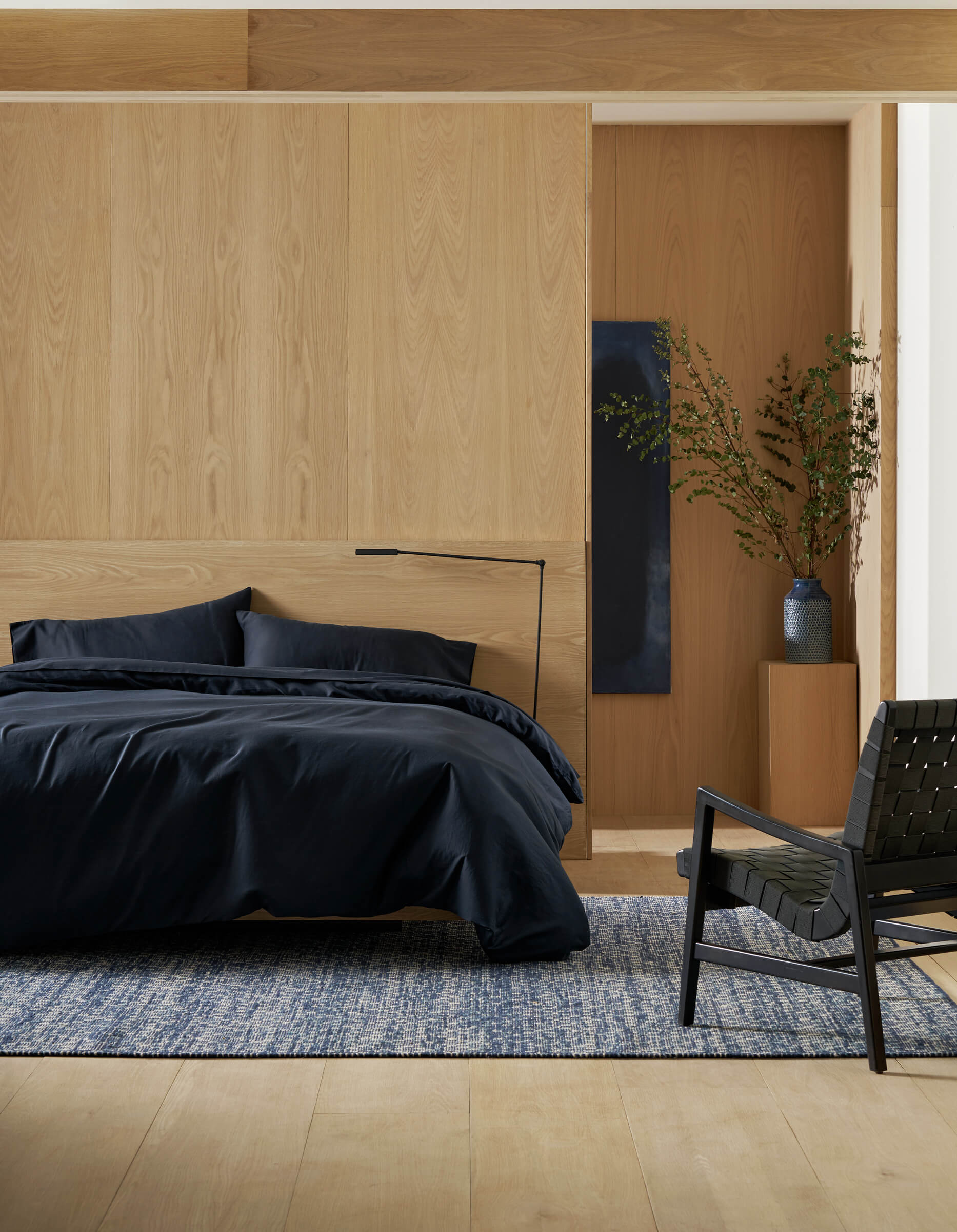 A modern bedroom with a wooden accent wall features the Soft-Wash Cotton Bedding Deluxe Bundle by Cozy Earth on the bed, a black floor lamp, a black woven chair, a blue rug, and a tall vase with branches on a wooden pedestal.