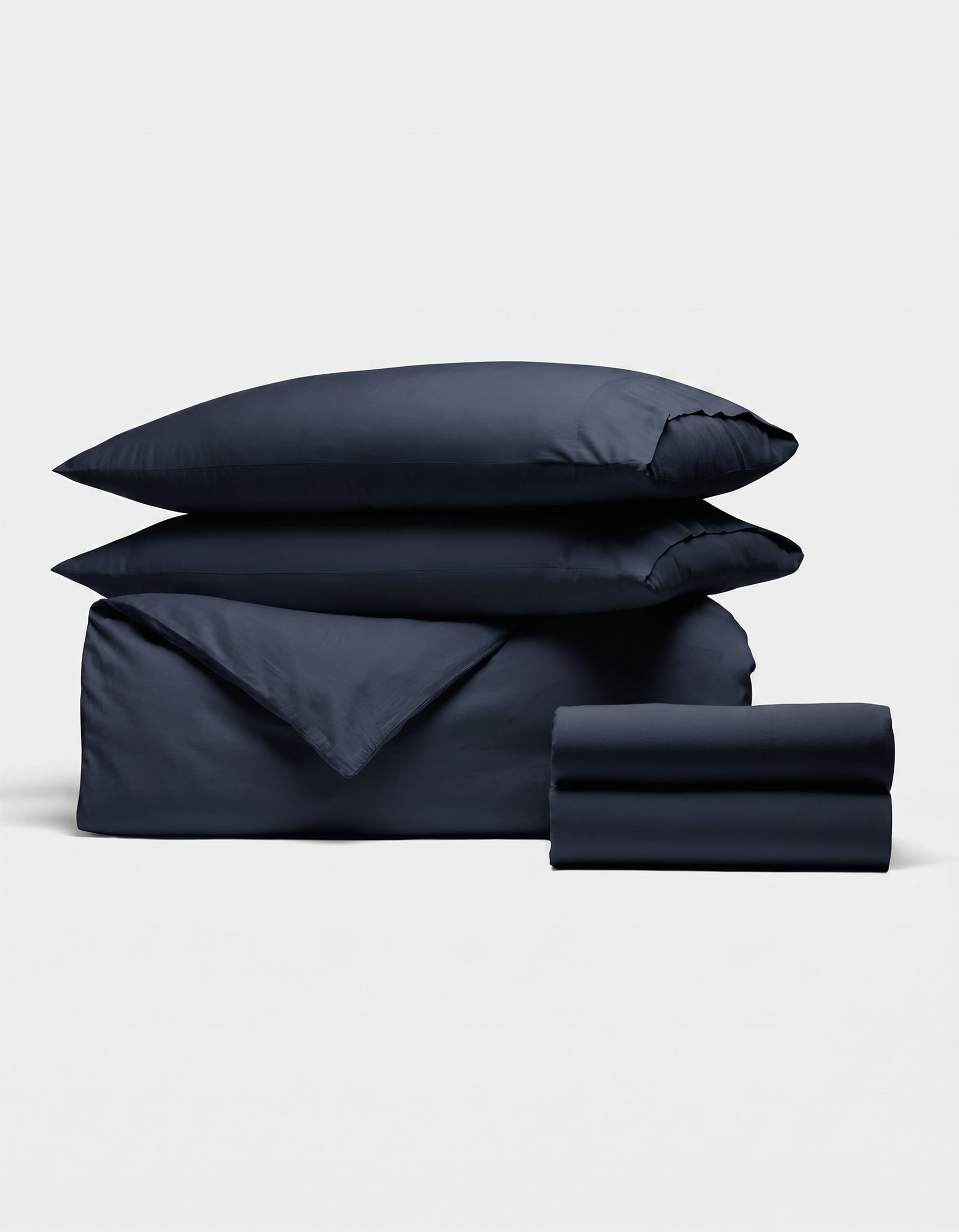 The Soft-Wash Cotton Bedding Deluxe Bundle by Cozy Earth features two pillows, a folded duvet, and sheet set in dark navy blue, neatly stacked against a plain white background.