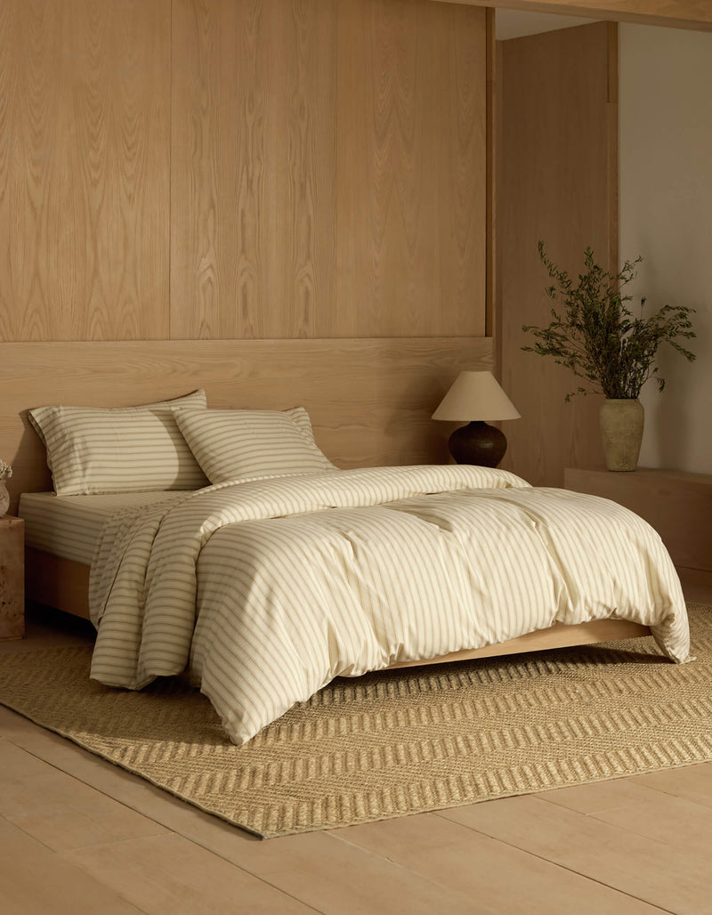 A neatly made bed with Cozy Earth's Soft-Wash Cotton Bedding Deluxe Bundle sits on a woven rug in a room with light wood walls and flooring. A nightstand with a lamp and vase of greenery stands beside the bed.
