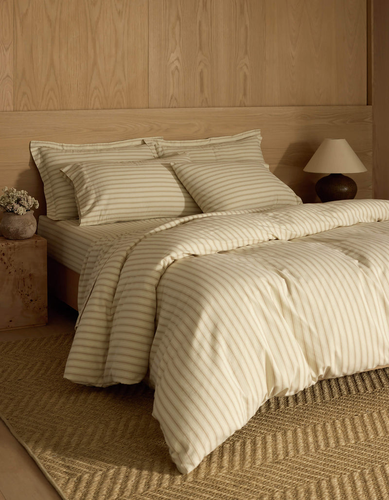 A neatly made bed with Cozy Earth's Soft-Wash Cotton Duvet Cover, matching pillowcases, and a wooden headboard sits against a light wood-paneled wall. A tan woven rug, side table with lamp, and vase of flowers complete the look.