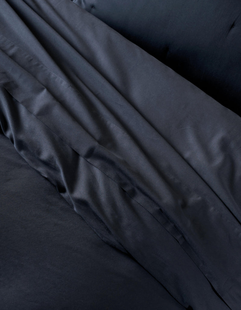 A close-up of the Cozy Earth Soft-Wash Cotton Duvet Cover shows neatly arranged black bedding and pillowcases, highlighting the smooth, slightly shiny fabric with gentle folds and creases.