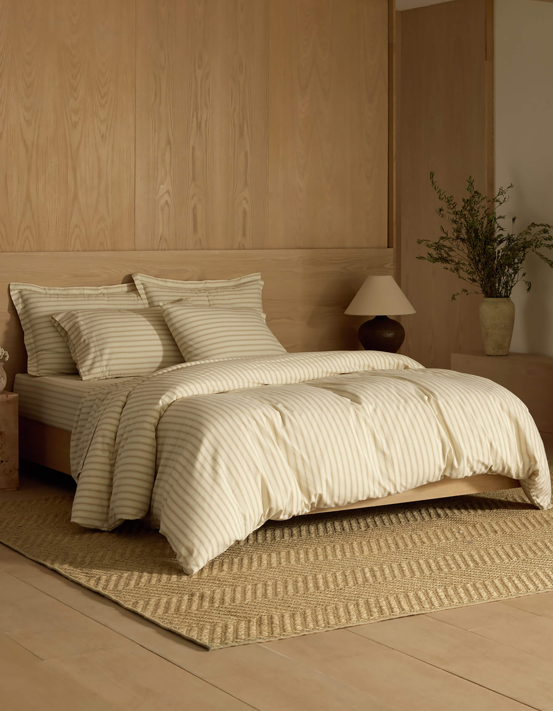 A minimalist bedroom features a neatly made bed dressed in the Cozy Earth Soft-Wash Cotton Duvet Cover, set atop a textured rug with light wood paneling, a nightstand, lamp, and a vase with greenery.