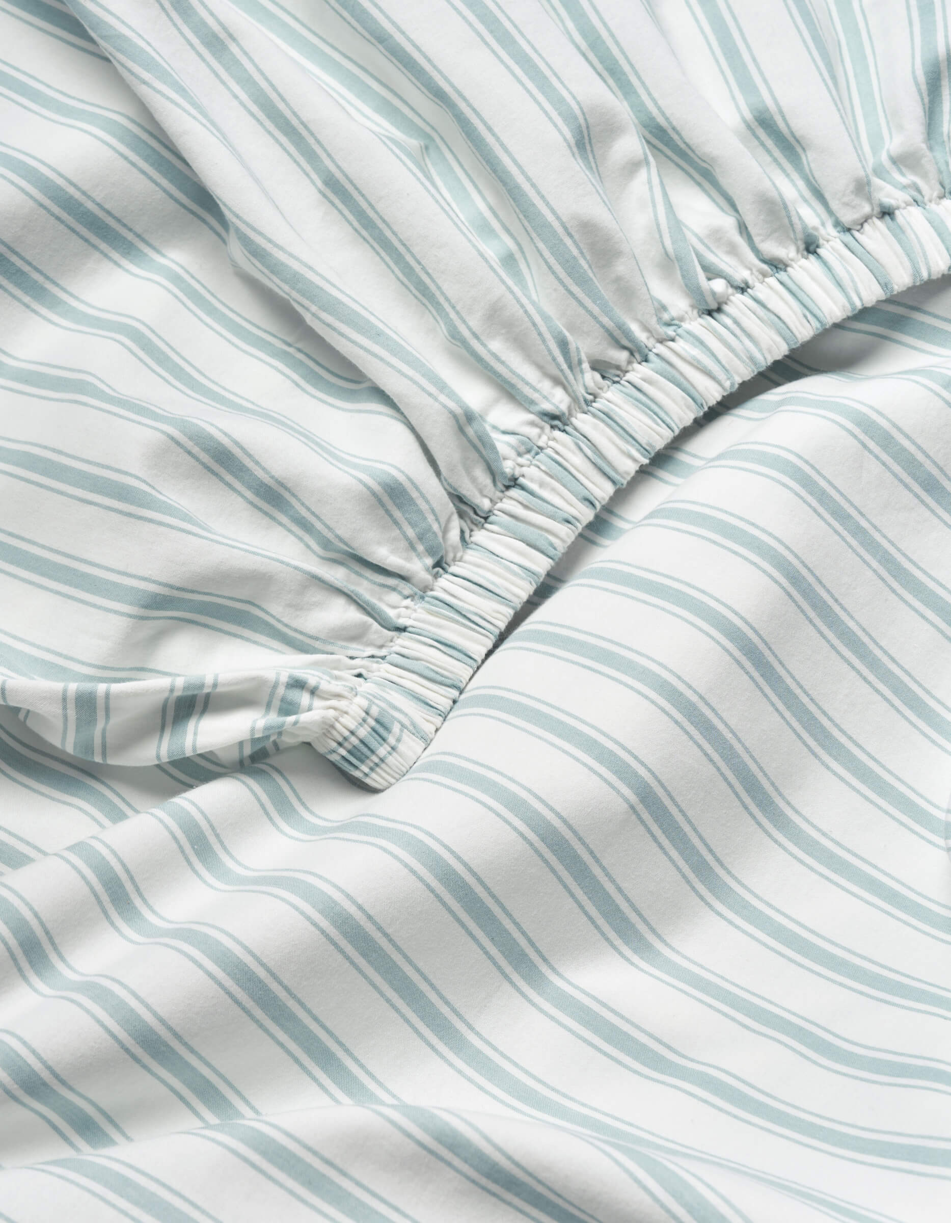 Close-up of the Soft-Wash Cotton Fitted Sheet by Cozy Earth, showing light blue and white vertical stripes and an elastic gathered edge for a snug fit.