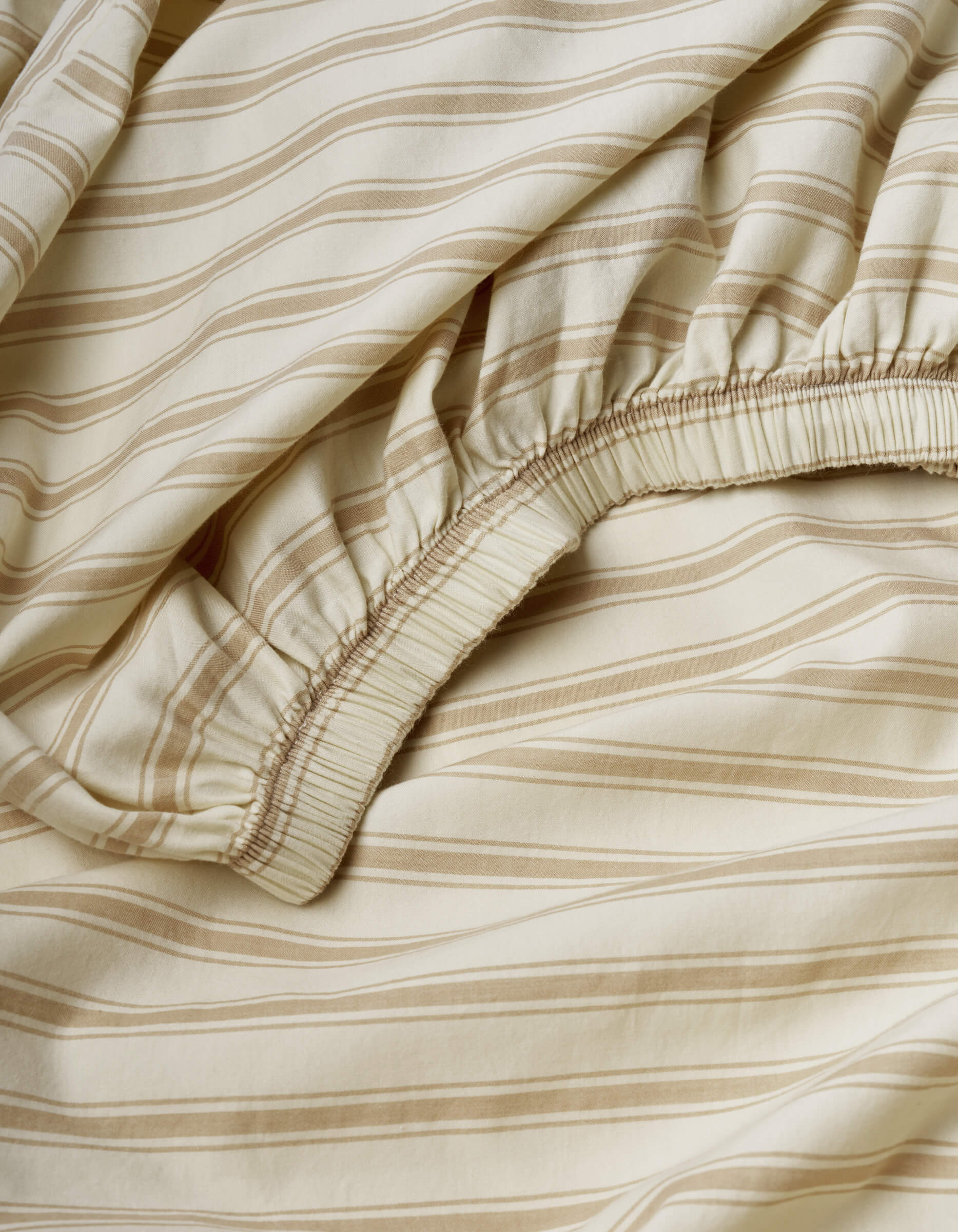 The Soft-Wash Cotton Fitted Sheet by Cozy Earth features a beige color with light brown horizontal stripes and an elastic edge, showcasing its gathered fabric detail, texture, and pattern.|Color:Coconut and Sandalwood Stripe