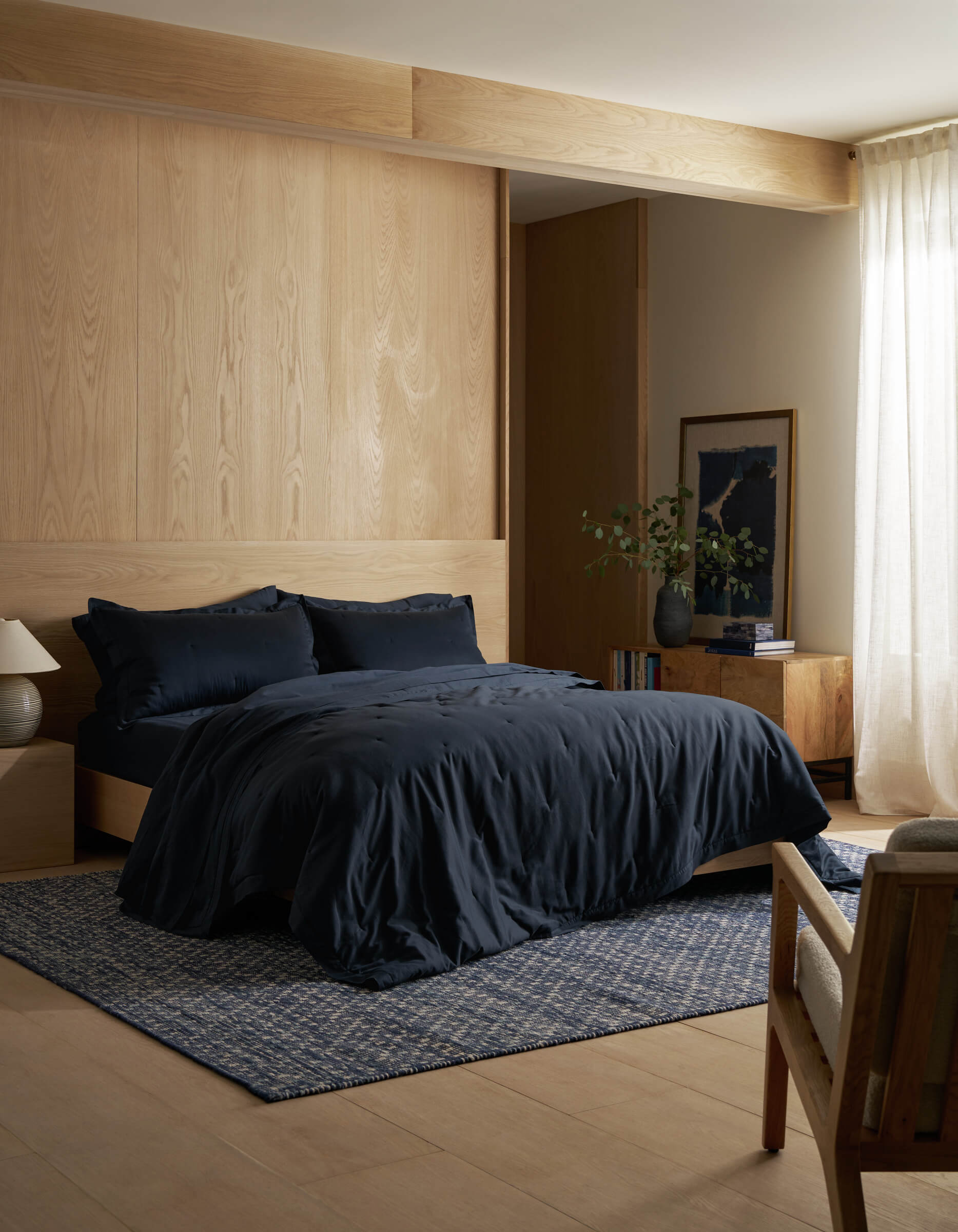 A modern bedroom features a wooden accent wall, a bed dressed in Cozy Earth’s Soft-Wash Cotton Quilt in dark blue, a small rug, wooden nightstand and dresser, a lamp, framed art, and sheer curtains allowing natural light.