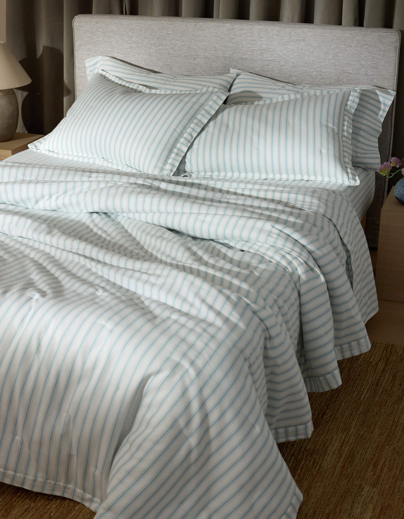 A neatly made bed features Cozy Earth's Soft-Wash Cotton Quilted Shams, with blue and white stripes, two pillows, and smooth, inviting bedding in a softly lit room with a light headboard, nightstand, and lamp.