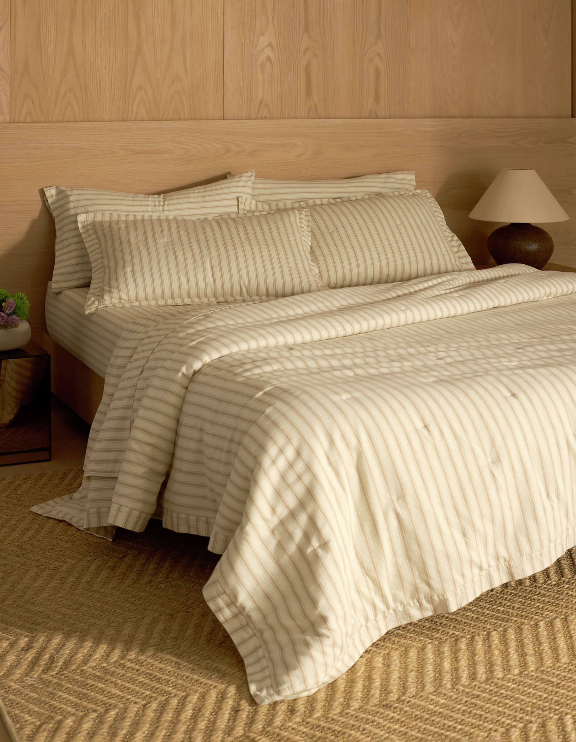 A bed featuring Cozy Earth's Soft-Wash Cotton Quilted Shams sits against a wooden paneled wall, complemented by matching pillows and a comforter. A bedside table with a lamp and vase, plus a woven rug on the floor, complete the cozy scene.