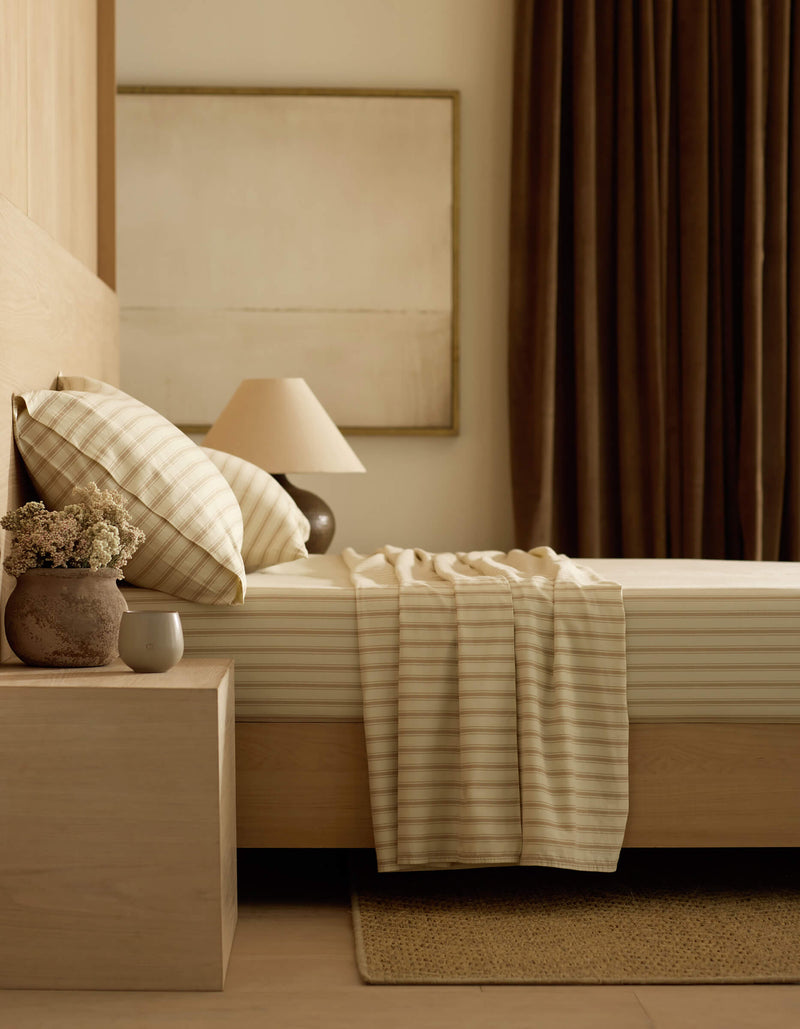 A minimalist bedroom features a light wood bed, nightstand, striped bedding with the Cozy Earth Soft-Wash Cotton Flat Sheet, a beige throw, table lamp, vase of flowers, ceramic cup, dark curtains, and a neutral abstract painting for a calm vibe.