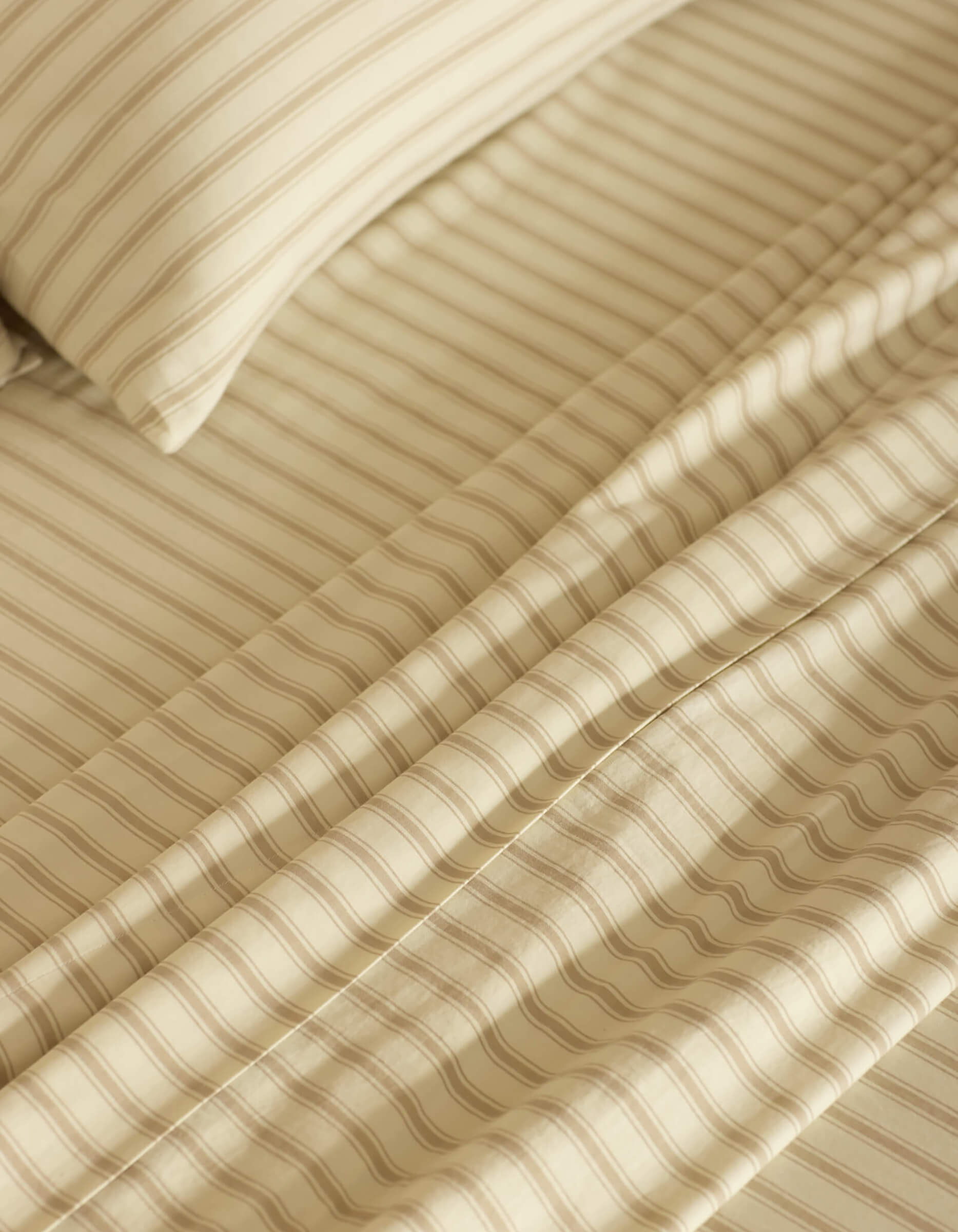 Close-up of a neatly made bed featuring the Cozy Earth Soft-Wash Cotton Sheet Set in beige and cream stripes. The smooth fabric and evenly spaced stripes create a calm, cozy atmosphere.|Color:Coconut and Sandalwood Stripe