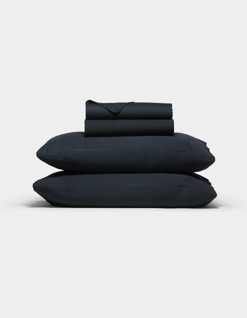 The Cozy Earth Soft-Wash Cotton Sheet Set, in dark navy blue, features two stacked pillows and folded sheets neatly arranged against a plain light gray background.