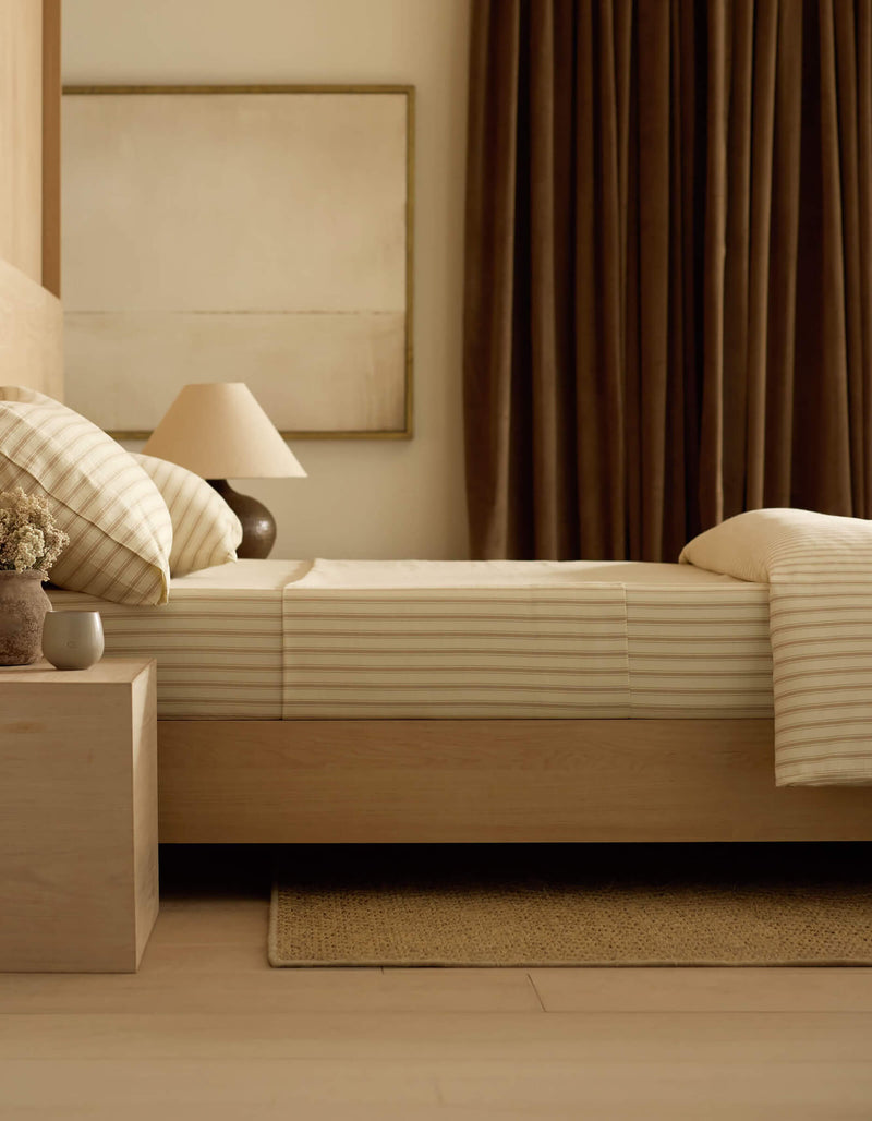 A neatly made bed with Cozy Earth’s Soft-Wash Cotton Sheet Set in beige and white stripes sits beside a wooden nightstand holding a lamp, vase of flowers, and cup in a softly lit, neutral-toned bedroom.