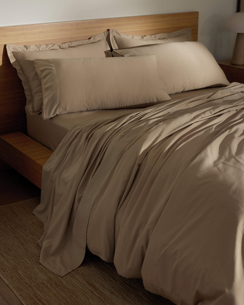 A neatly made bed features Cozy Earth's Soft-Wash Cotton Pillowcases in light brown, with matching sheets and comforter on a wooden frame. The bedding feels smooth and soft, complementing the room's minimal, warm-toned decor.