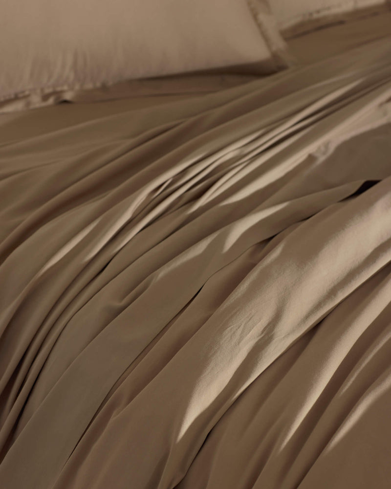 The Cozy Earth Soft-Wash Cotton Duvet Cover in brown is spread over the bed, creating gentle folds and shadows. Its smooth surface looks inviting, with matching pillowcases partially visible in the background.