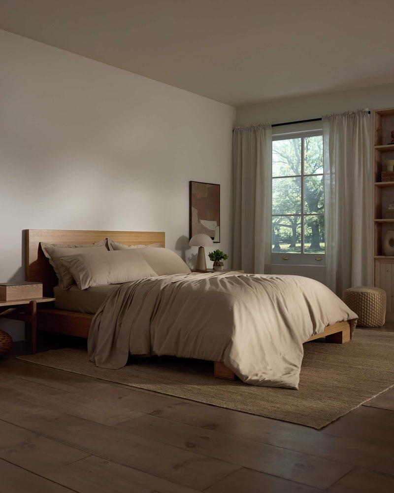 A cozy bedroom featuring a wooden bed frame with Cozy Earth's Soft-Wash Cotton Duvet Cover, a nightstand and lamp, sheer curtains allowing natural light, neutral decor, and wooden flooring.