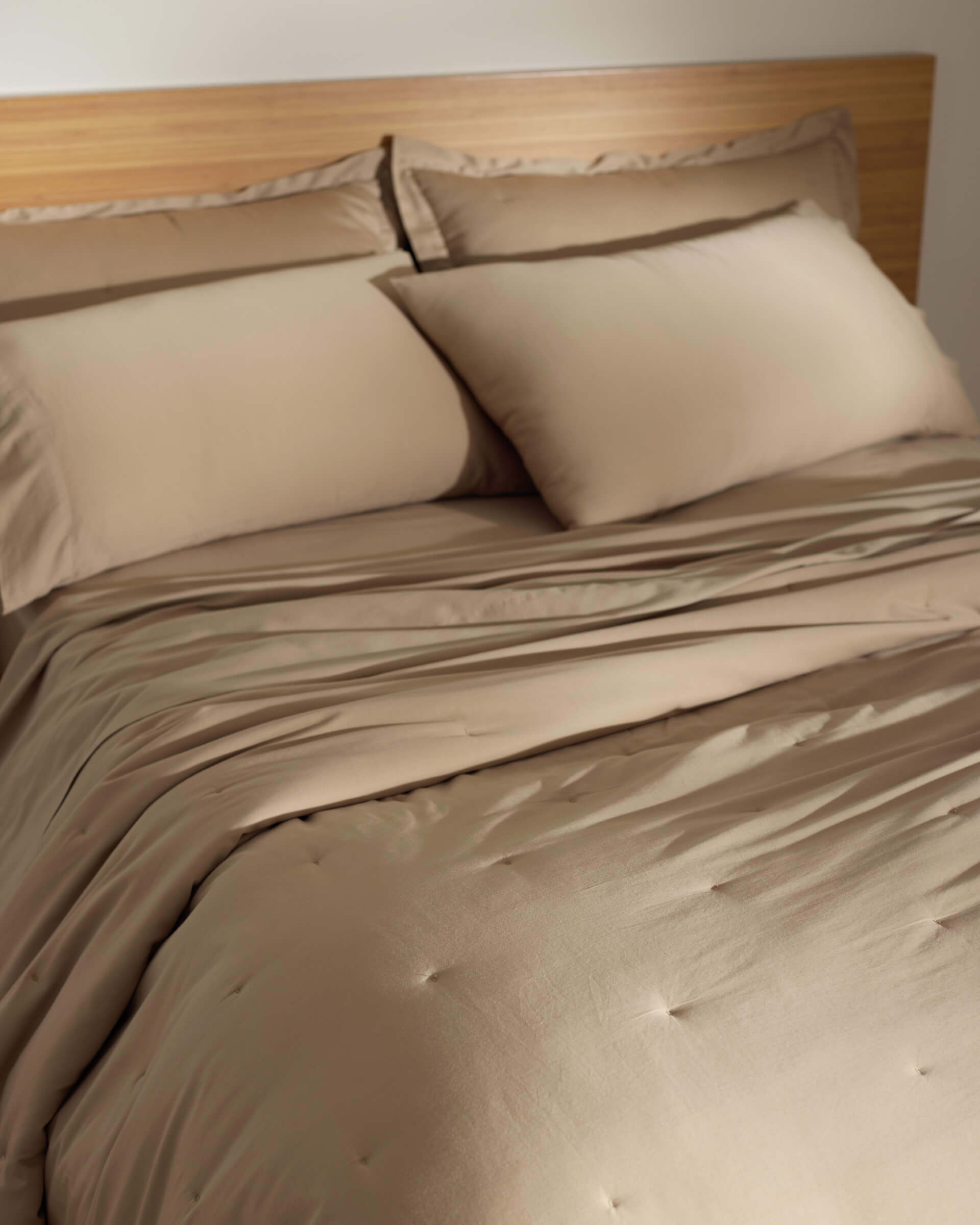 A neatly made bed with the Cozy Earth Soft-Wash Cotton Quilt, featuring beige sheets and pillowcases, is set against a wooden headboard in a softly lit room.