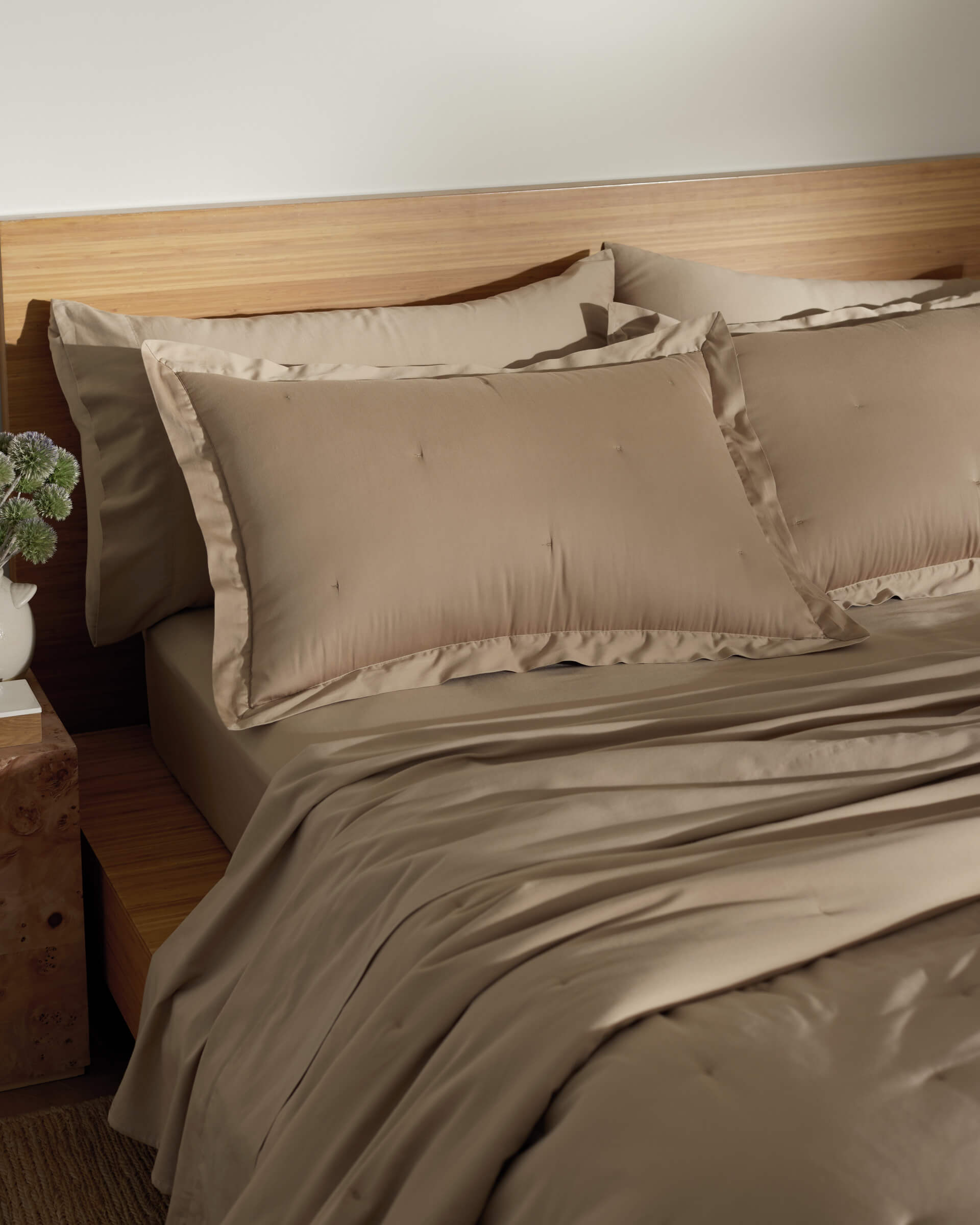 A neatly made bed features Cozy Earth's Soft-Wash Cotton Quilted Shams in beige, paired with matching sheets and a duvet against a wooden headboard. A small nightstand with a decorative plant completes the calm and cozy ambiance.