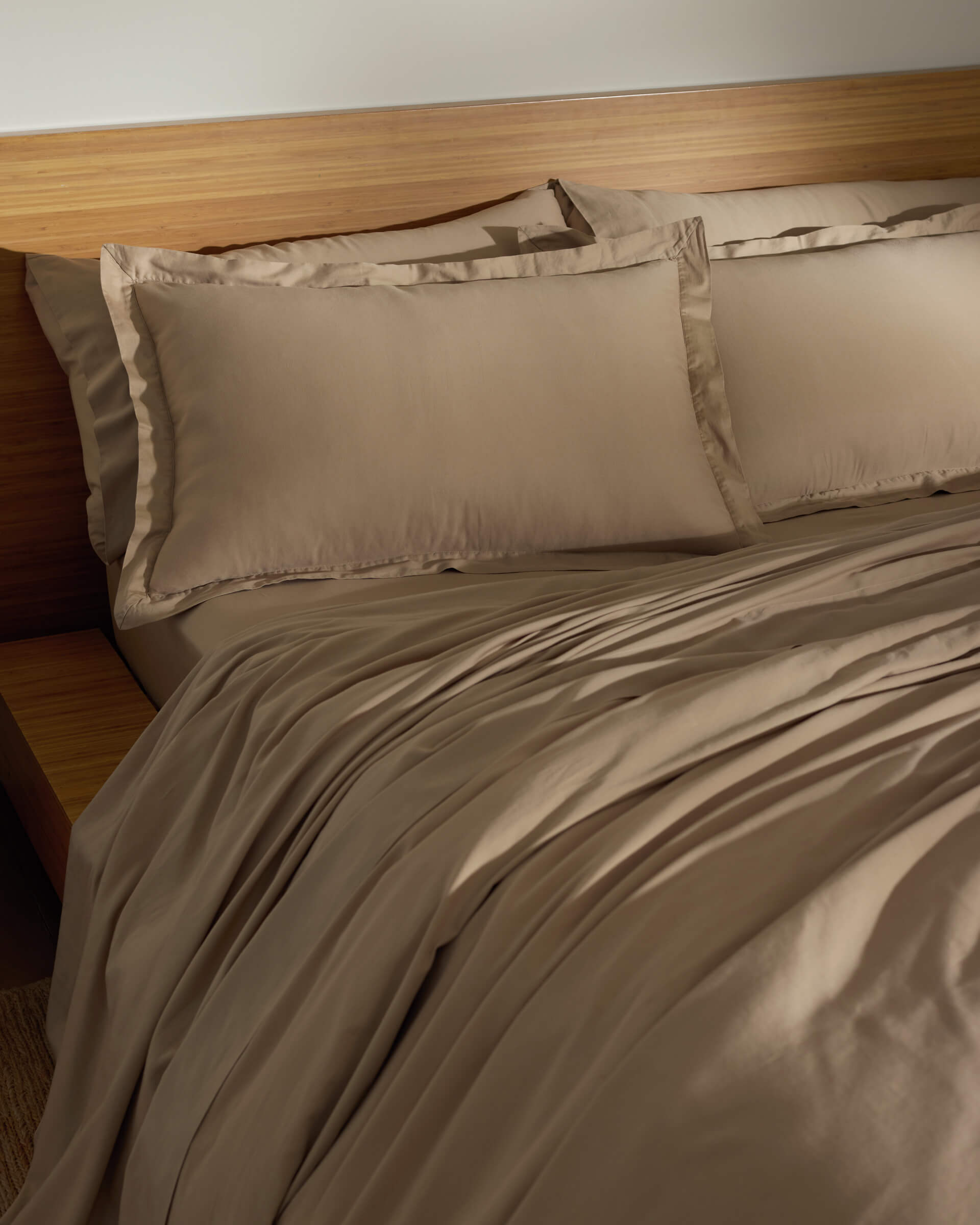 A softly lit room features a neatly made bed with the Cozy Earth Soft-Wash Cotton Duvet Set—light brown sheets, pillowcases, and matching comforter—set against a wooden headboard and side table.|Color:Sandalwood