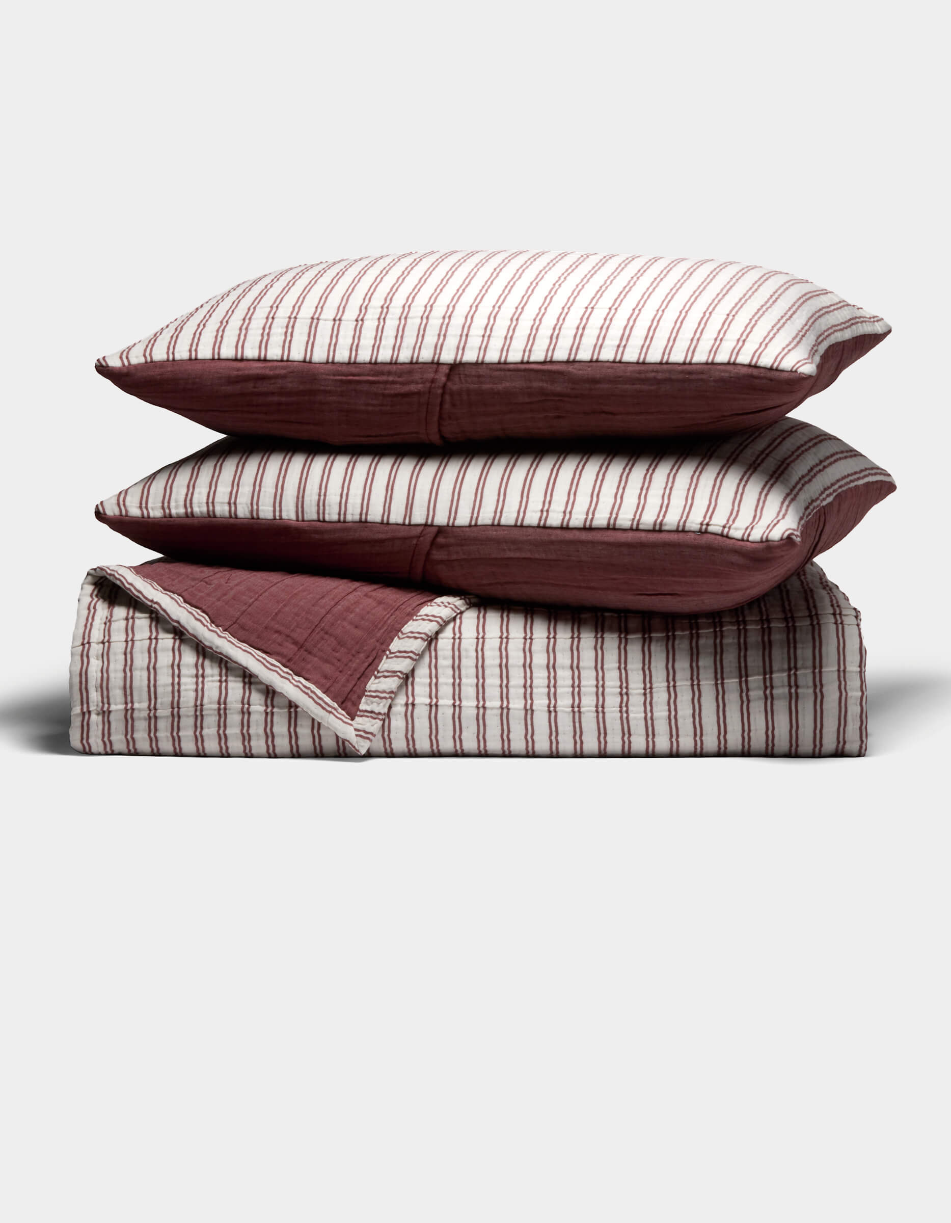 The Waterfront Coverlet Bundle by Cozy Earth features two pillows with red and white vertical stripes stacked on a matching folded quilt with a solid red underside, all displayed against a plain light background.|Color:Currant Stripe