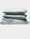 The Cozy Earth Waterfront Coverlet Bundle shows two pillows and a folded quilt stacked neatly, each with blue and white stripes on one side and solid blue on the other, all set against a plain white background.|Color:River Stripe