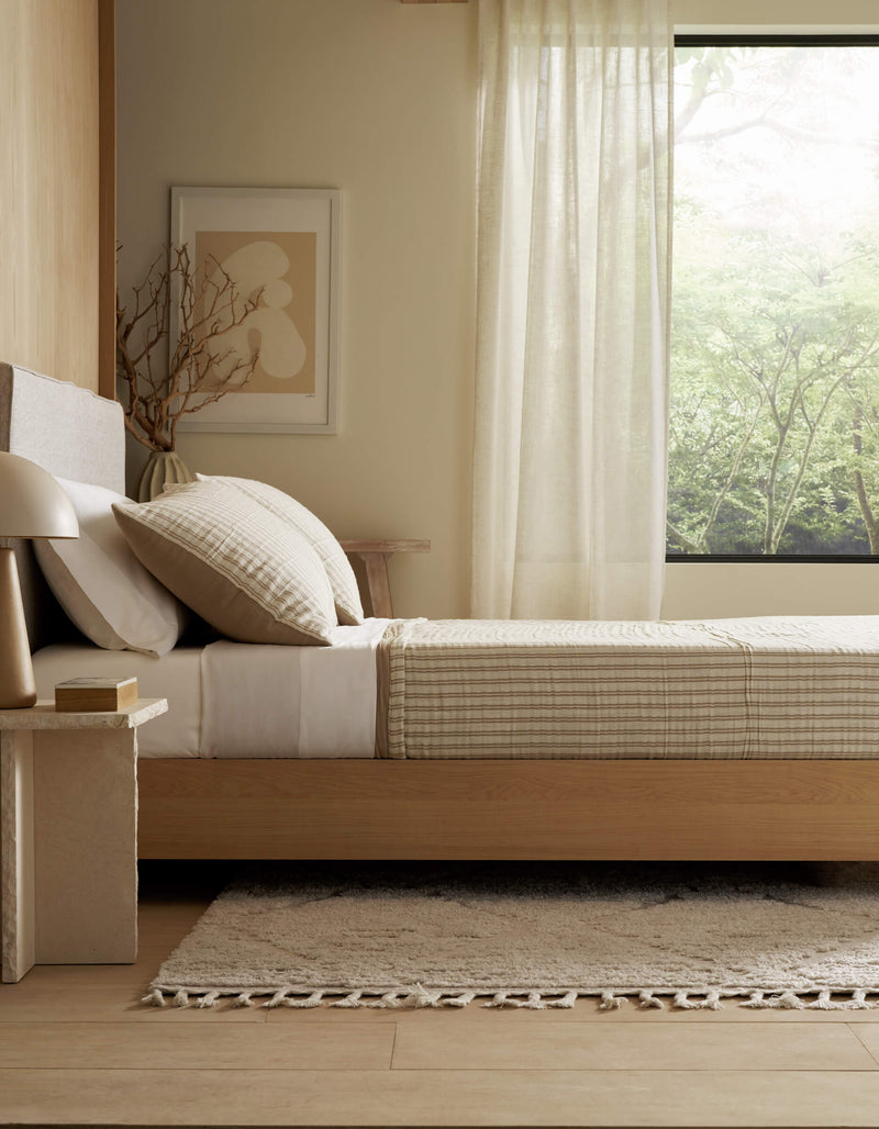 A minimalist bedroom featuring Cozy Earth's Waterfront Coverlet Bundle, a wooden bed, nightstand with lamp and book, abstract wall art, and a large window with sheer curtains overlooking greenery.